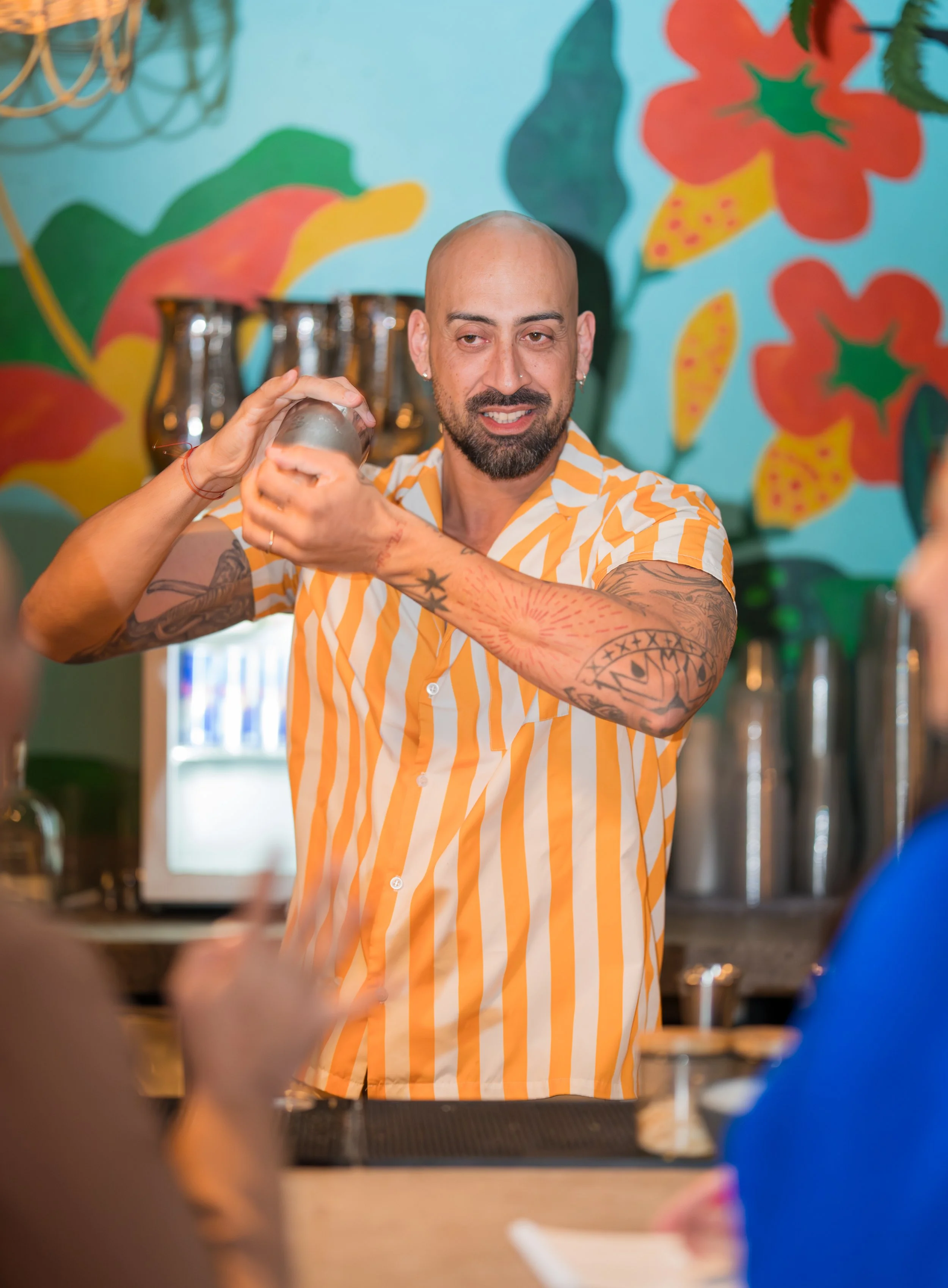 Bartender making a cocktail at a bar with colorful floral mural in background, wearing a yellow and white striped shirt, with tattoos on his arms.