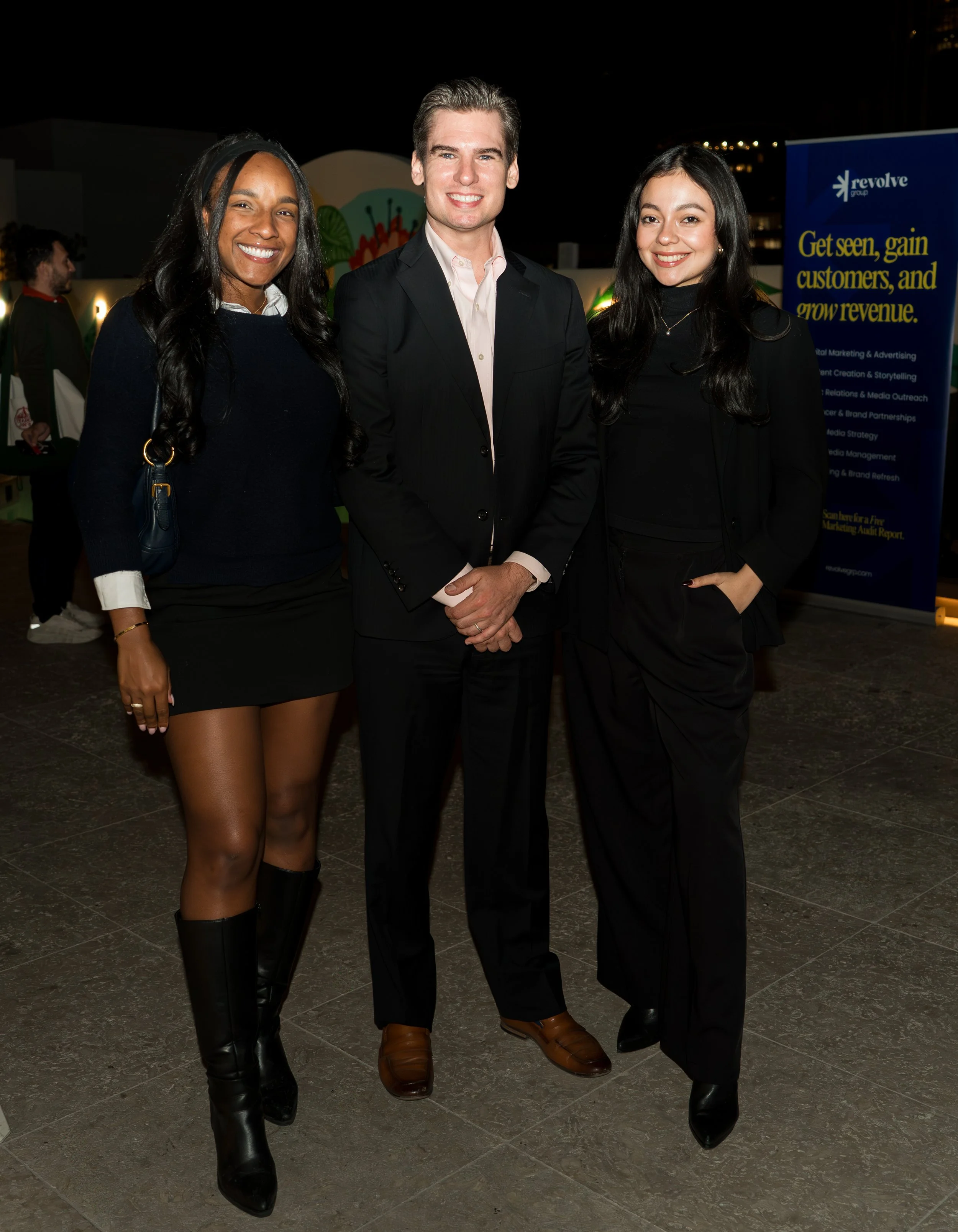 Three people, two women and one man, standing together at a professional event, smiling at the camera. The woman on the left is wearing a black skirt, black boots, and a navy sweater, while the woman on the right is dressed in black, and the man in t