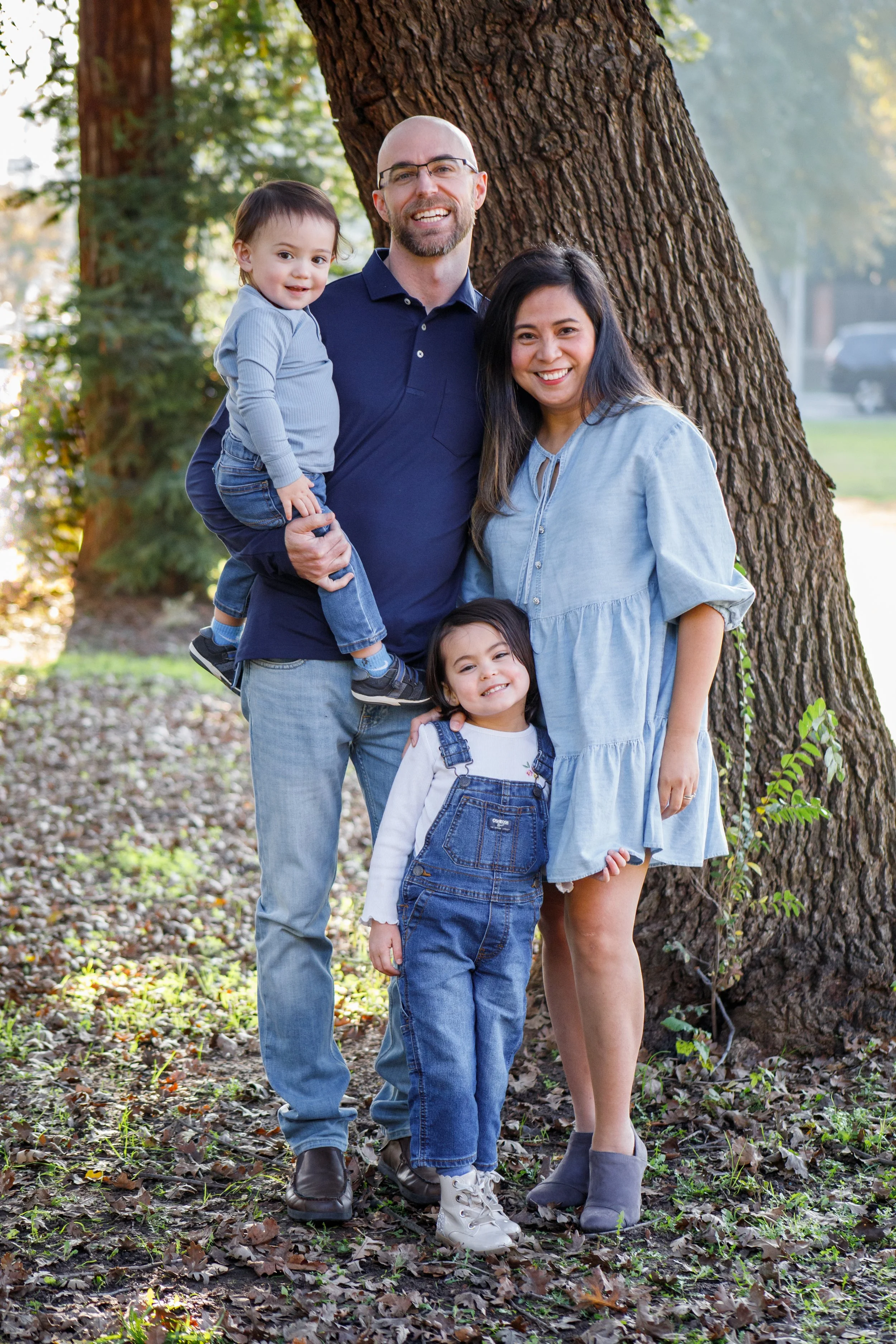A family of four standing outdoors near a large tree, smiling, with sunlight filtering through the leaves.