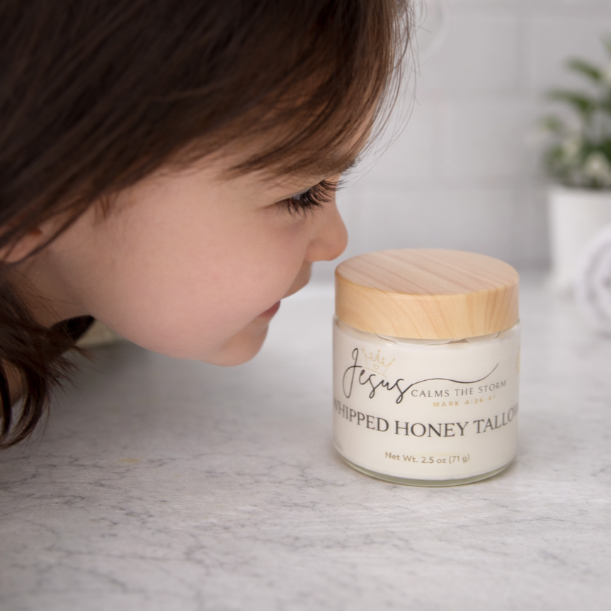 Close-up of a young girl smelling a jar of whipped honey tallow on a white countertop with a potted plant in the background.