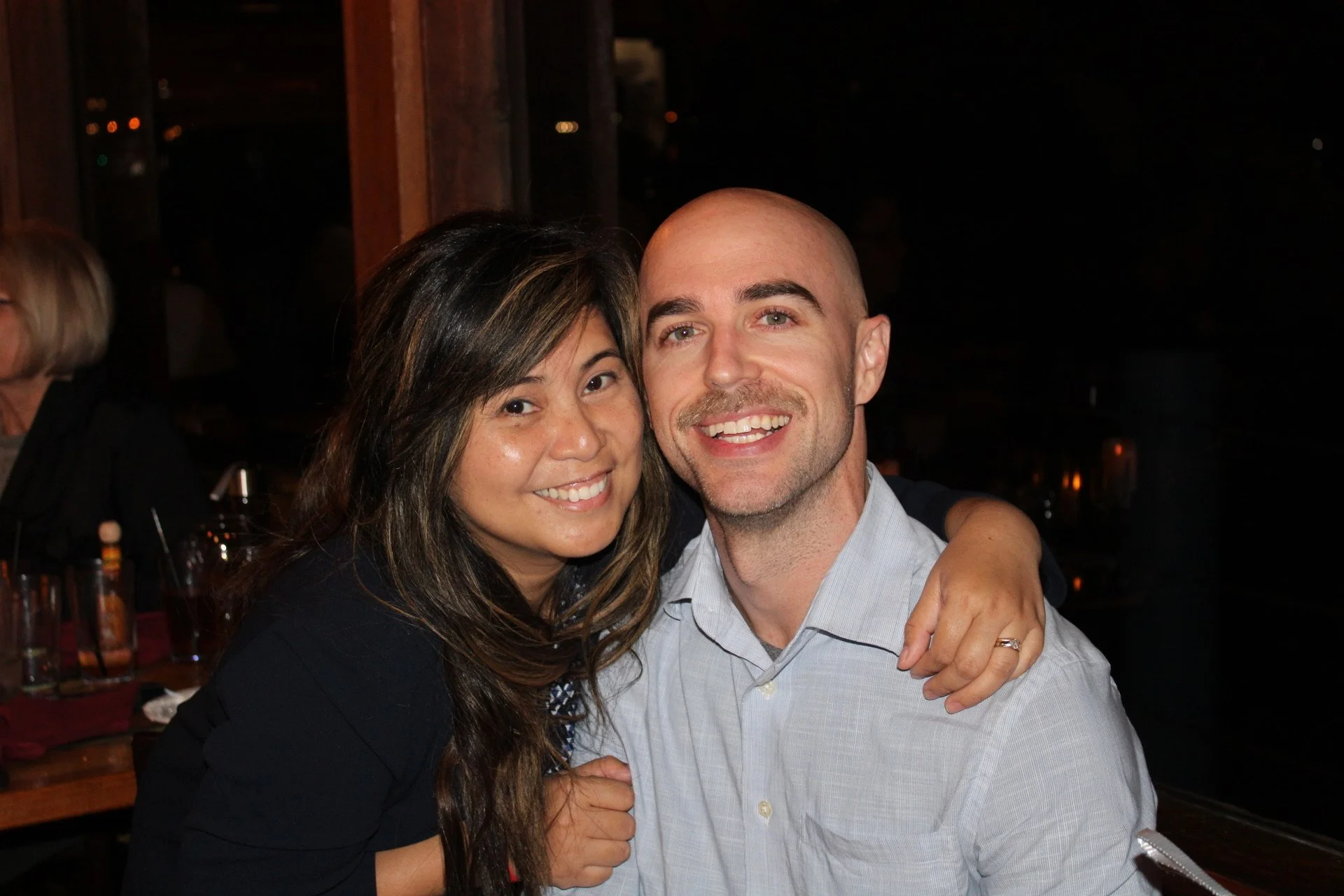 A smiling woman and man sitting together at a restaurant or bar, with the woman embracing the man, in a warm and happy setting.