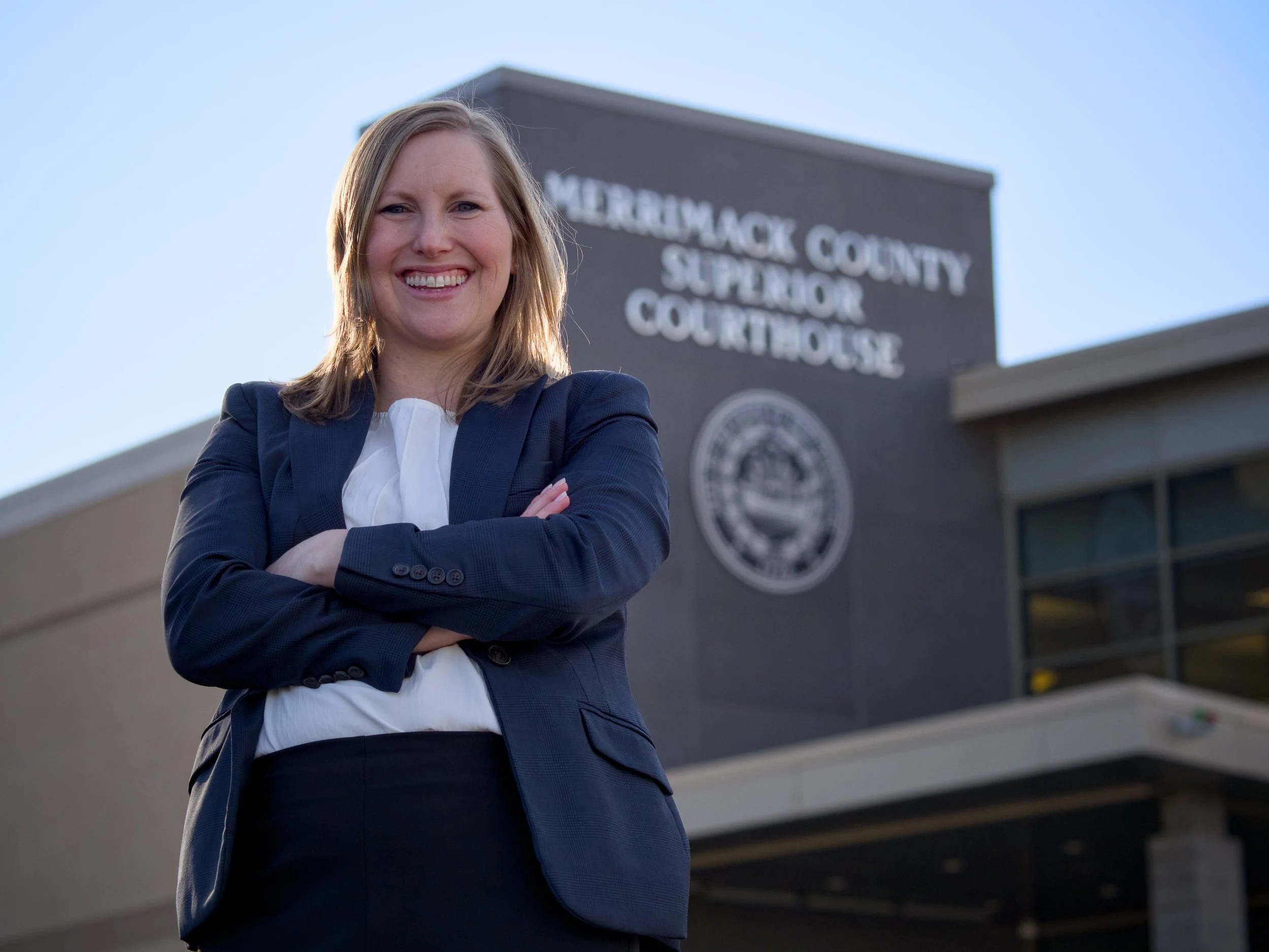A woman named Meg Hagaman with a smile, wearing a navy blazer and white shirt, stands outside the Merrimack County Superior Courthouse with her arms crossed.