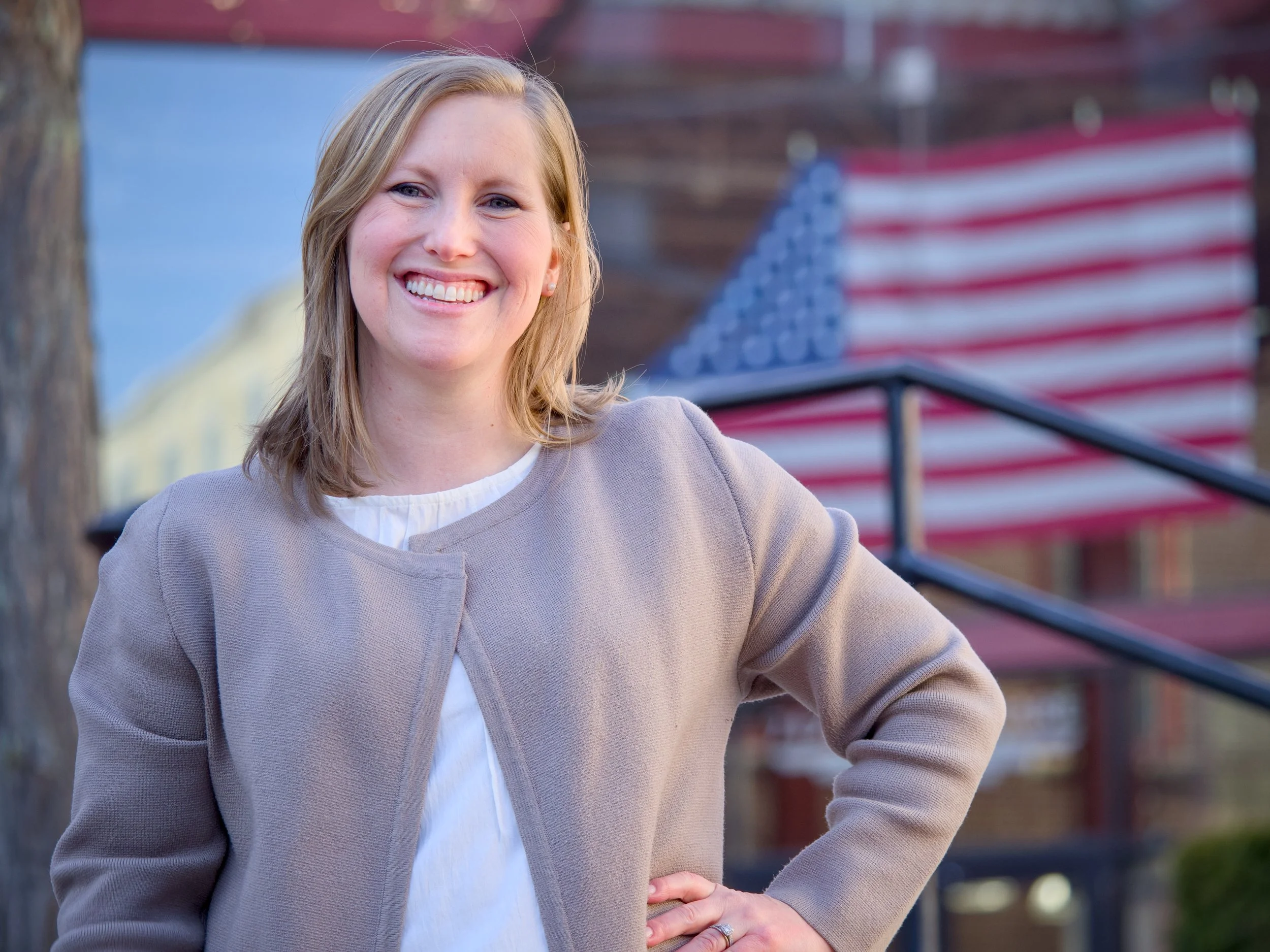 A smiling woman named Meg Hagaman with blonde hair standing outdoors near an American flag in a city setting.