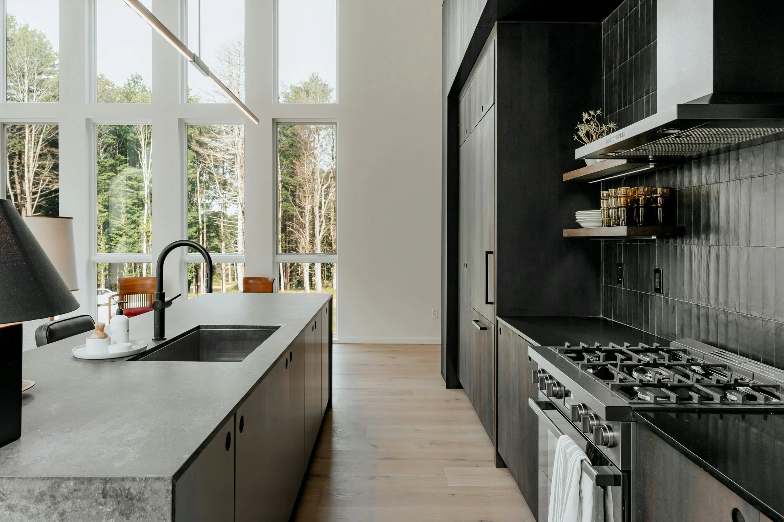 Modern kitchen with large windows, black cabinetry, black backsplash, and stainless steel appliances.