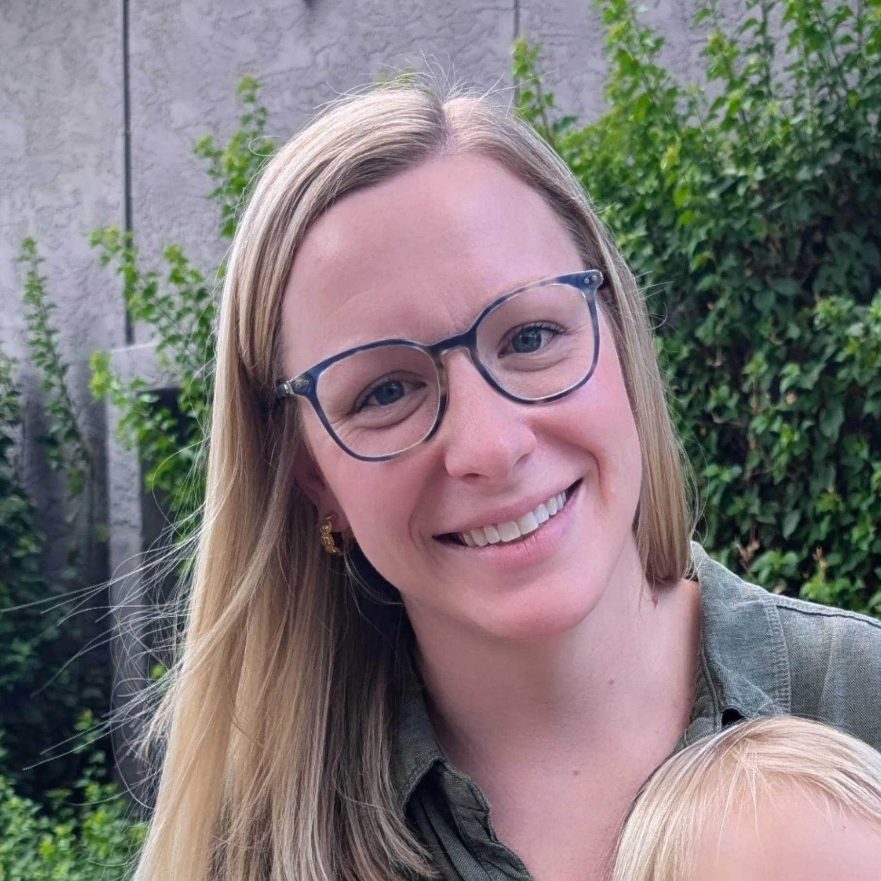 A woman with long blonde hair wearing glasses and earrings, smiling outdoors with green foliage and a gray wall in the background.