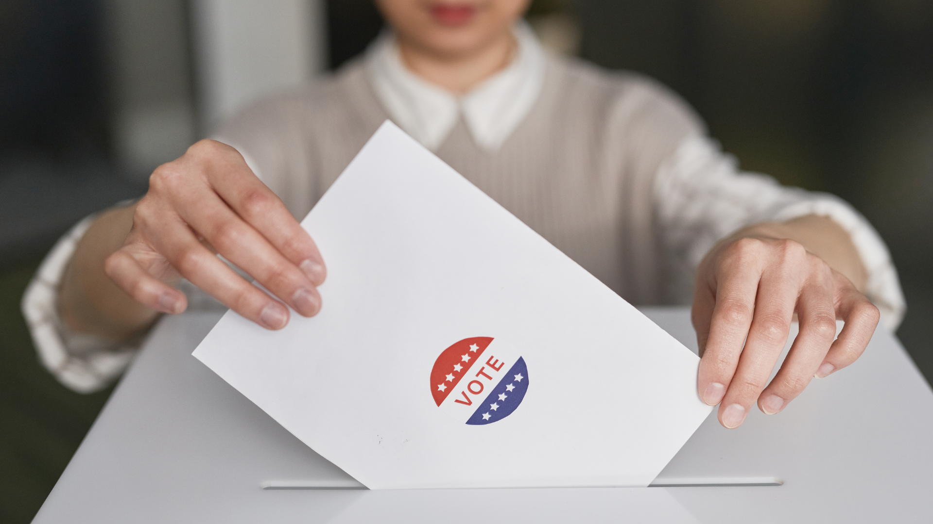 Person placing a ballot with a 'VOTE' sticker into a ballot box.