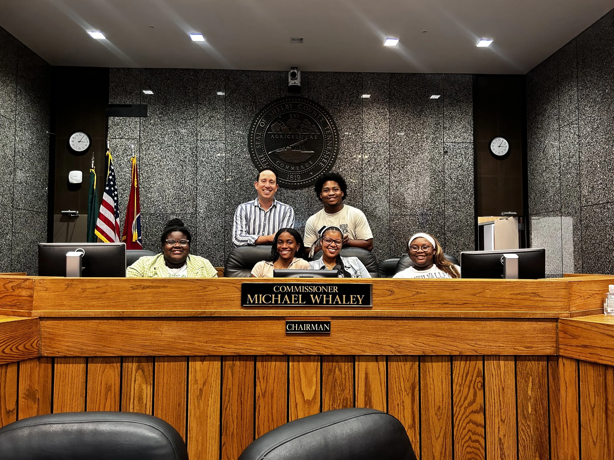 Group of five people in a conference room, with three sitting at a wooden desk and two standing behind them. The desk has a sign that reads "Commissioner Michael Whaley" and "Chairman." The background features the seal of Shelby County, flags, and clocks on the wall.