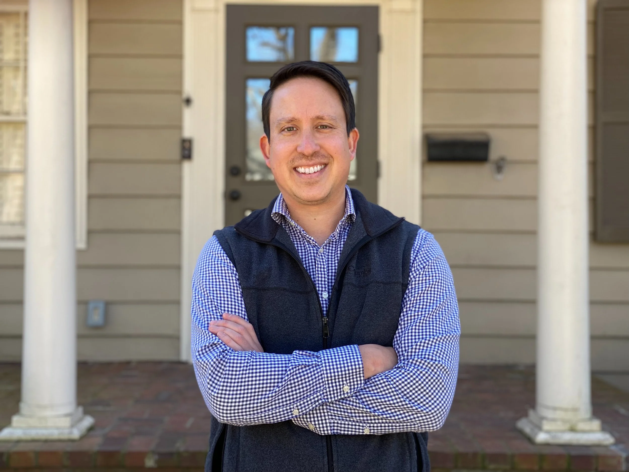 A man standing outside a house, smiling with arms crossed, wearing a checked shirt and a vest.
