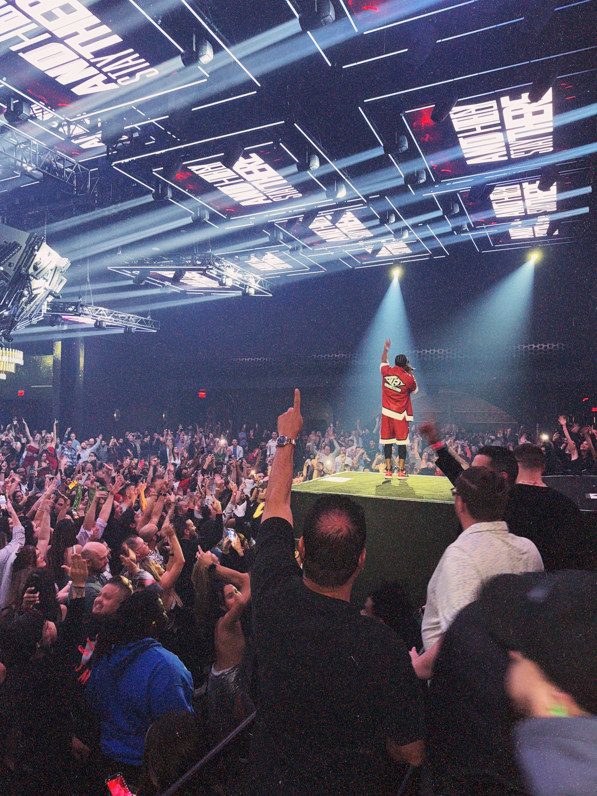 Ludacris on stage with a microphone, wearing a red jacket and black shorts, wearing a hat, standing under a spotlight, in a large crowd at a concert or party with many people raising their hands.