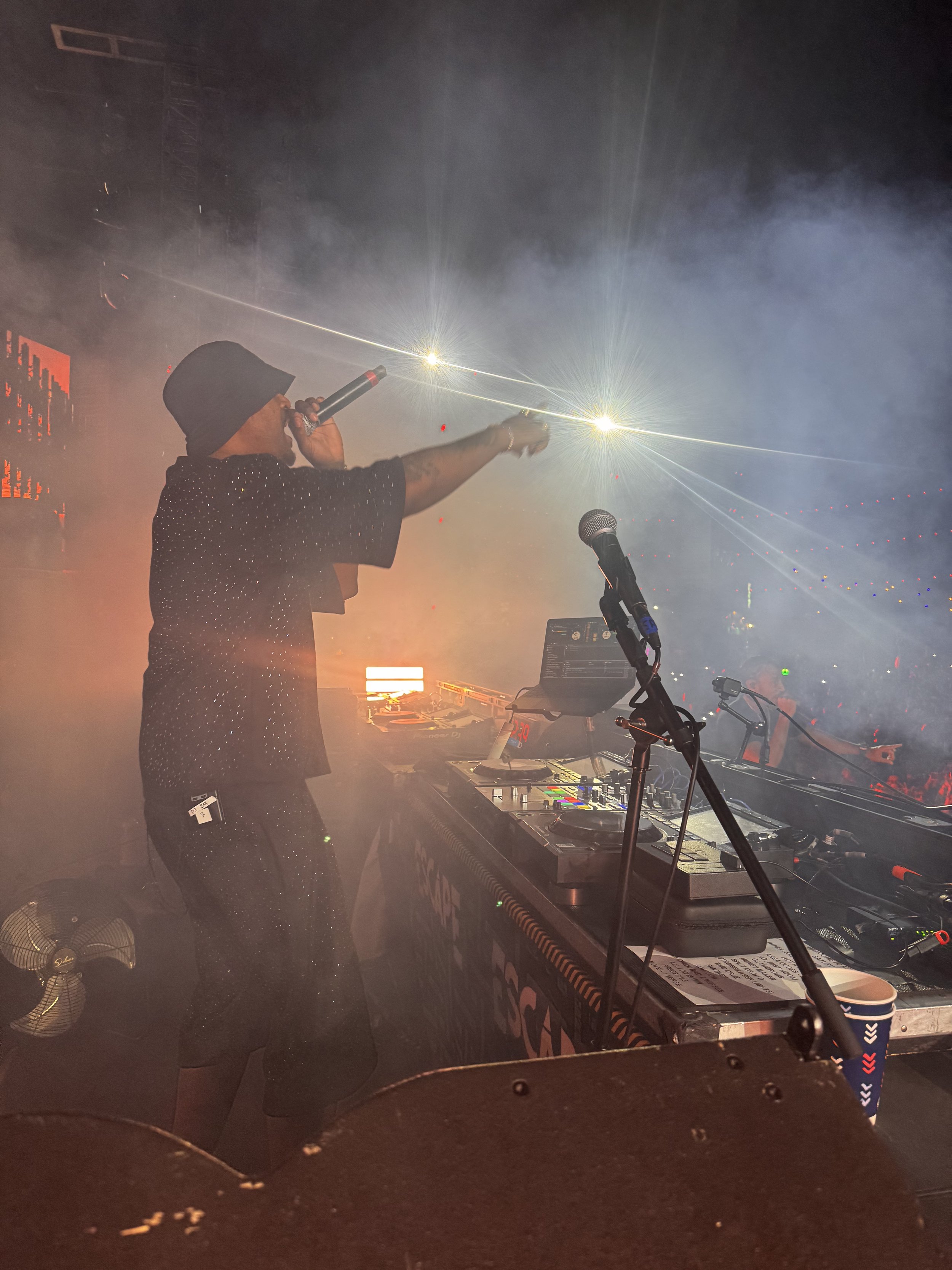 A DJ or performer on stage in a dark, smoky setting, wearing a black hat and dotted black shirt, holding a microphone in one hand and raising the other arm. The stage has DJ equipment, including a laptop and turntables, with a microphone on a stand and a paper with notes. Bright lights and special effects are visible in the background.