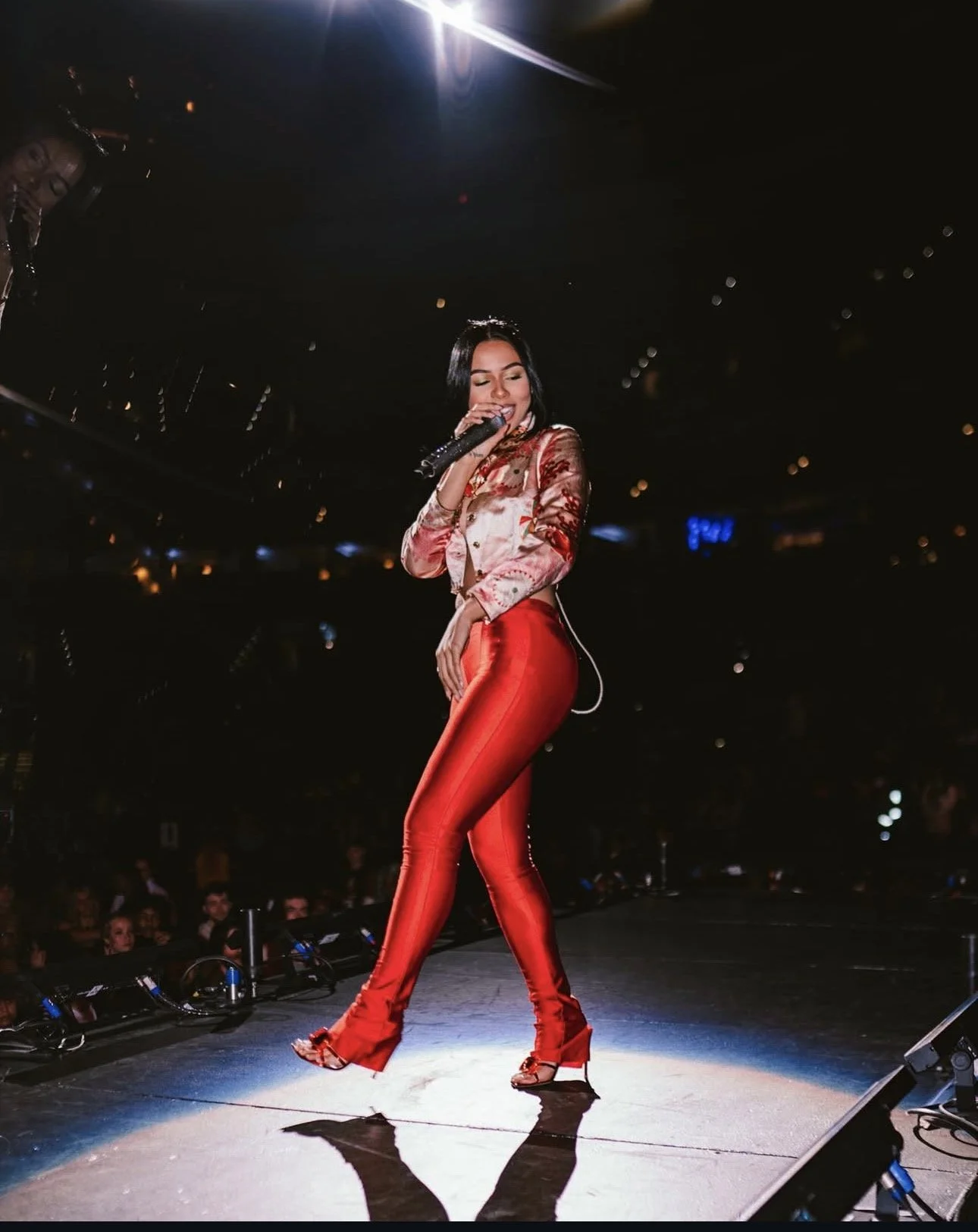 A female singer performing on stage, wearing a shiny red pants and a silk blazer with floral patterns, holding a microphone and smiling. The setting is dark with stage lights.