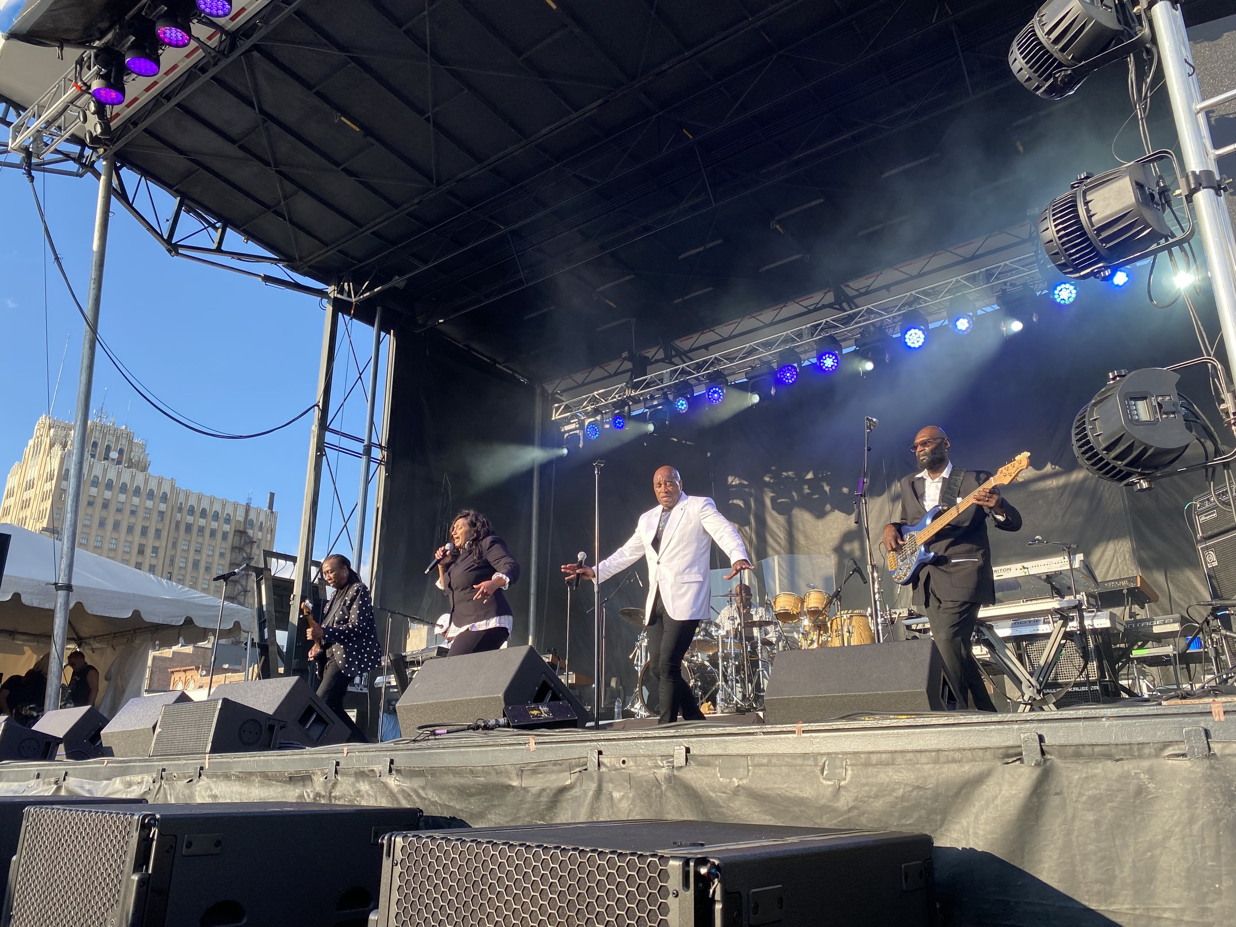 A live outdoor concert with five musicians on stage, including a female singer, a male singer in a white jacket, and a bass guitarist in a black suit, with a cityscape and blue sky in the background.