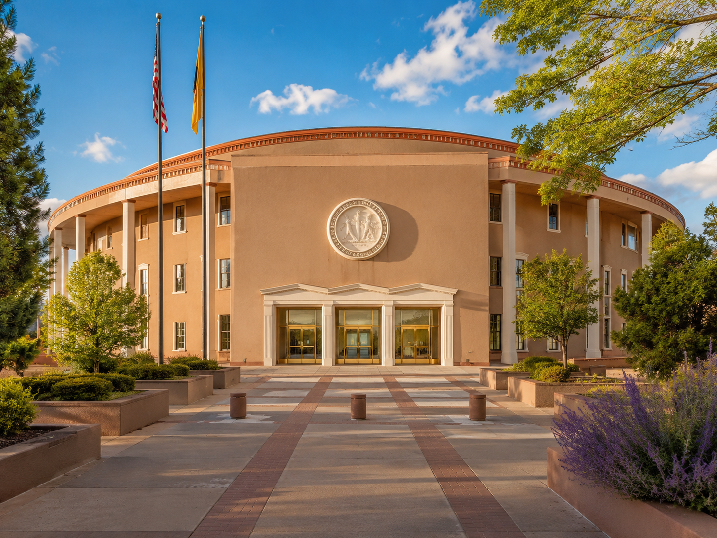 A government or civic building with a symmetrical entrance flanked by large white columns, a circular seal or emblem on the wall above the entrance, and two flags on tall flagpoles in front. The building is surrounded by trees and landscaped flower beds, with a paved walkway leading to the entrance. The sky is clear with a few clouds.