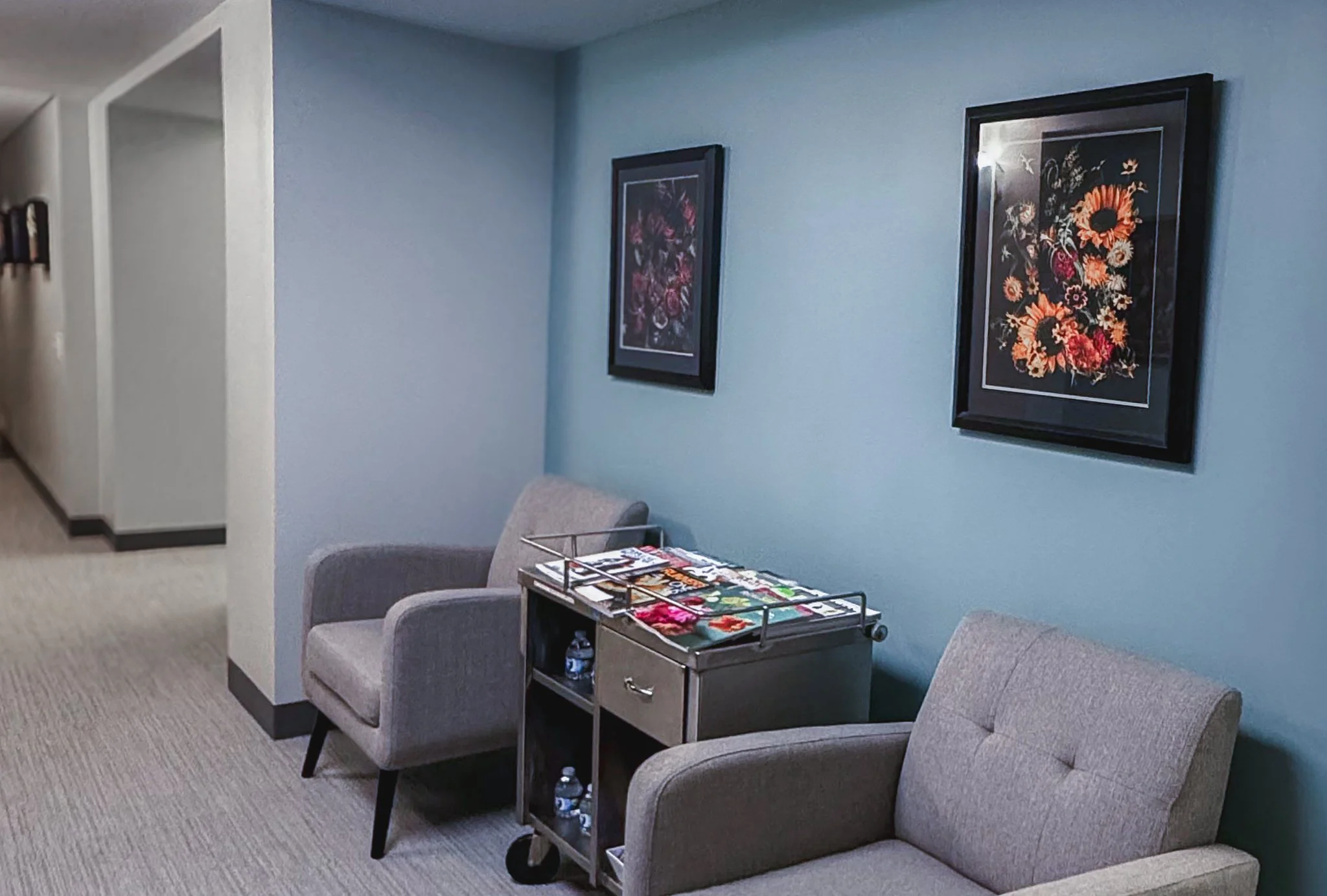 A cozy waiting or living room area with a light green wall, two framed floral art prints, a beige armchair, a small wooden side table with magazines and water bottles, and a carpeted floor.