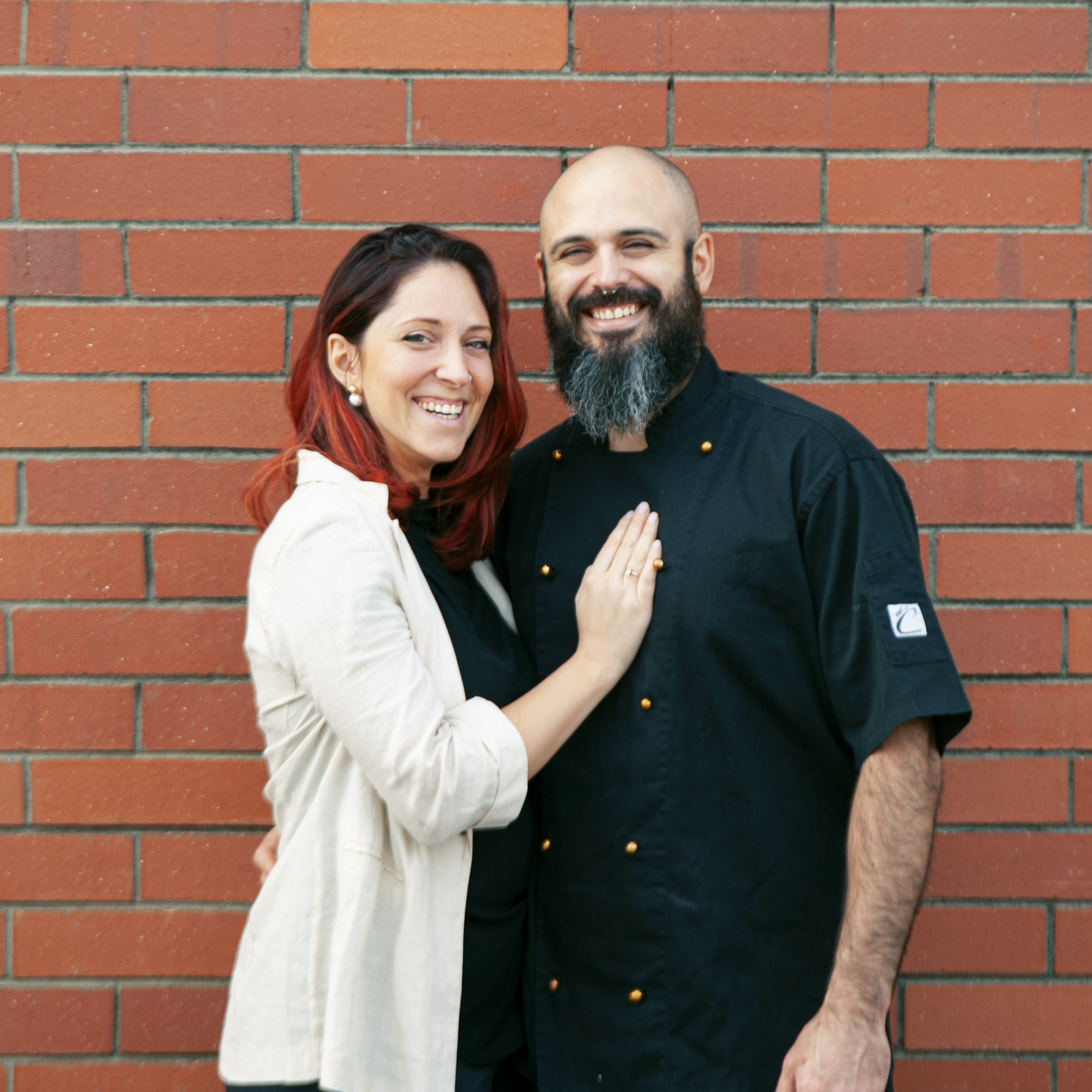 A woman with red hair and a pearl earring smiling next to a bald man with a beard and a big smile, standing against a red brick wall.