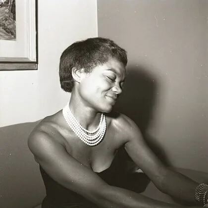Black-and-white photograph of a woman with short hair, wearing a strapless dress and pearl necklaces, smiling with her eyes closed.