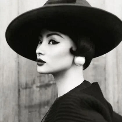 A woman wearing a large wide-brimmed hat and vintage-style earrings, facing sideways in black and white.