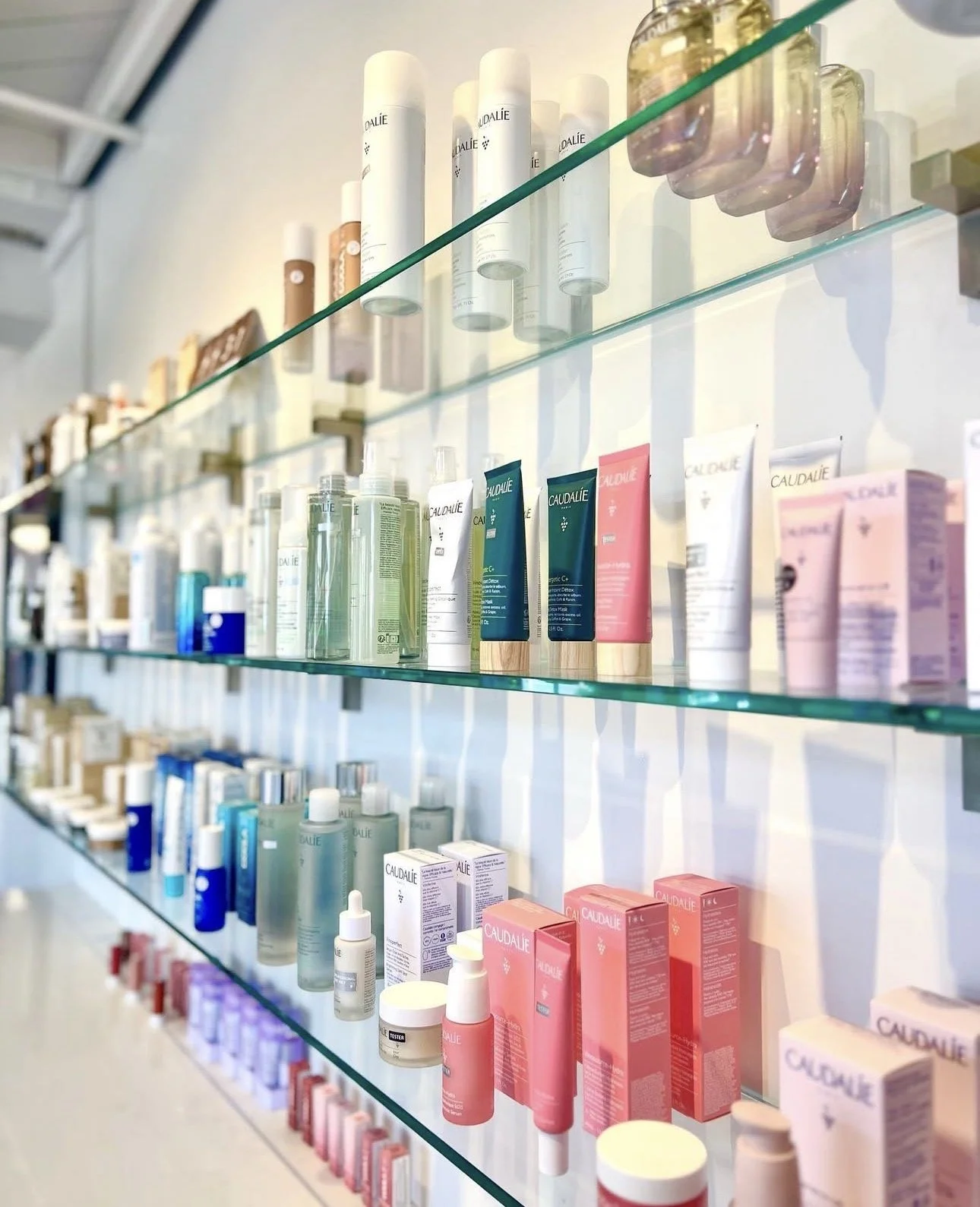 Glass shelves on a wall holding various skincare products, bottles, and tubes, mainly from the brand Caudalie.