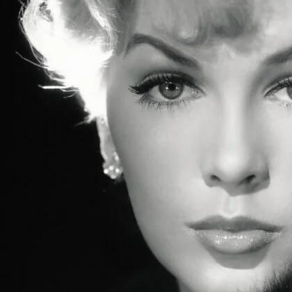 Close-up black and white photograph of a woman with short wavy hair and makeup, with one eye slightly squinting.