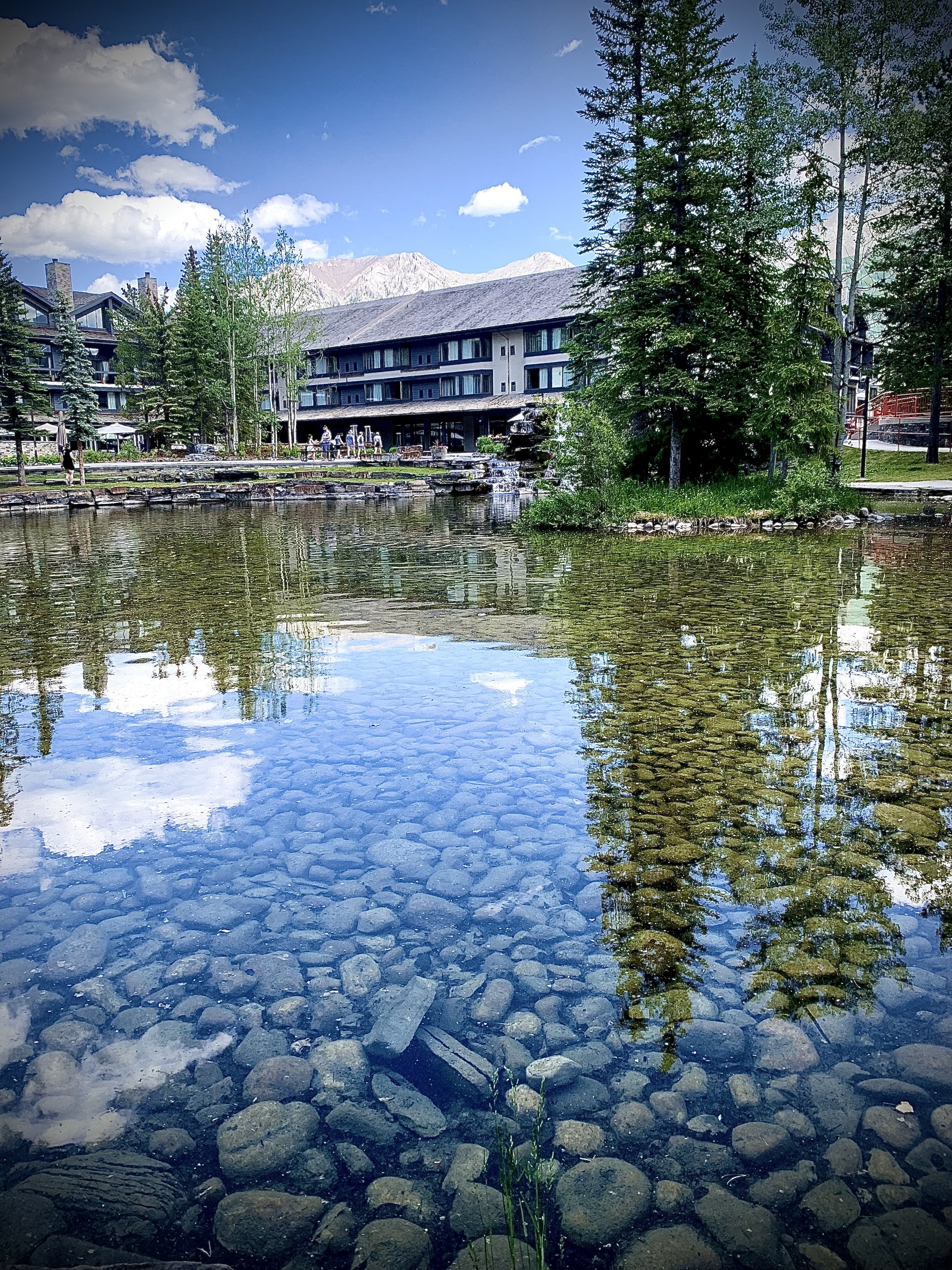 A clear pond with visible rocks and reflections of trees and the sky, with a building and mountain in the background.