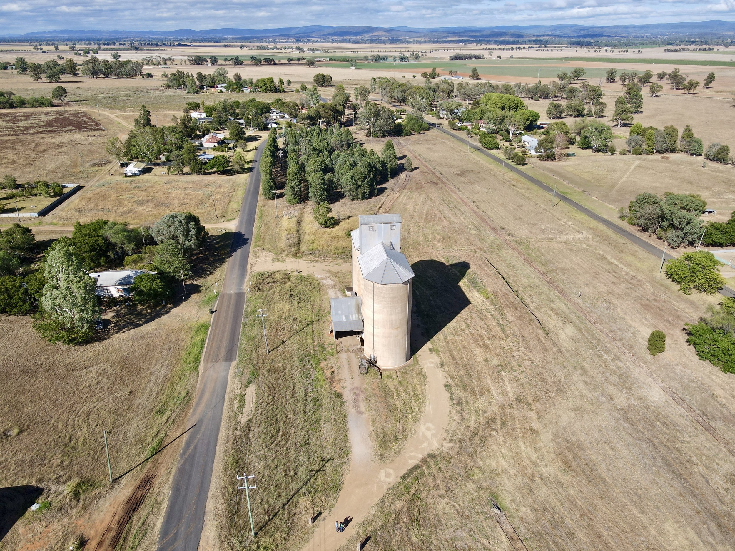 Aerial view of a rural landscape with a grain silo, farmhouses, trees, open fields, and a road in a farming area.