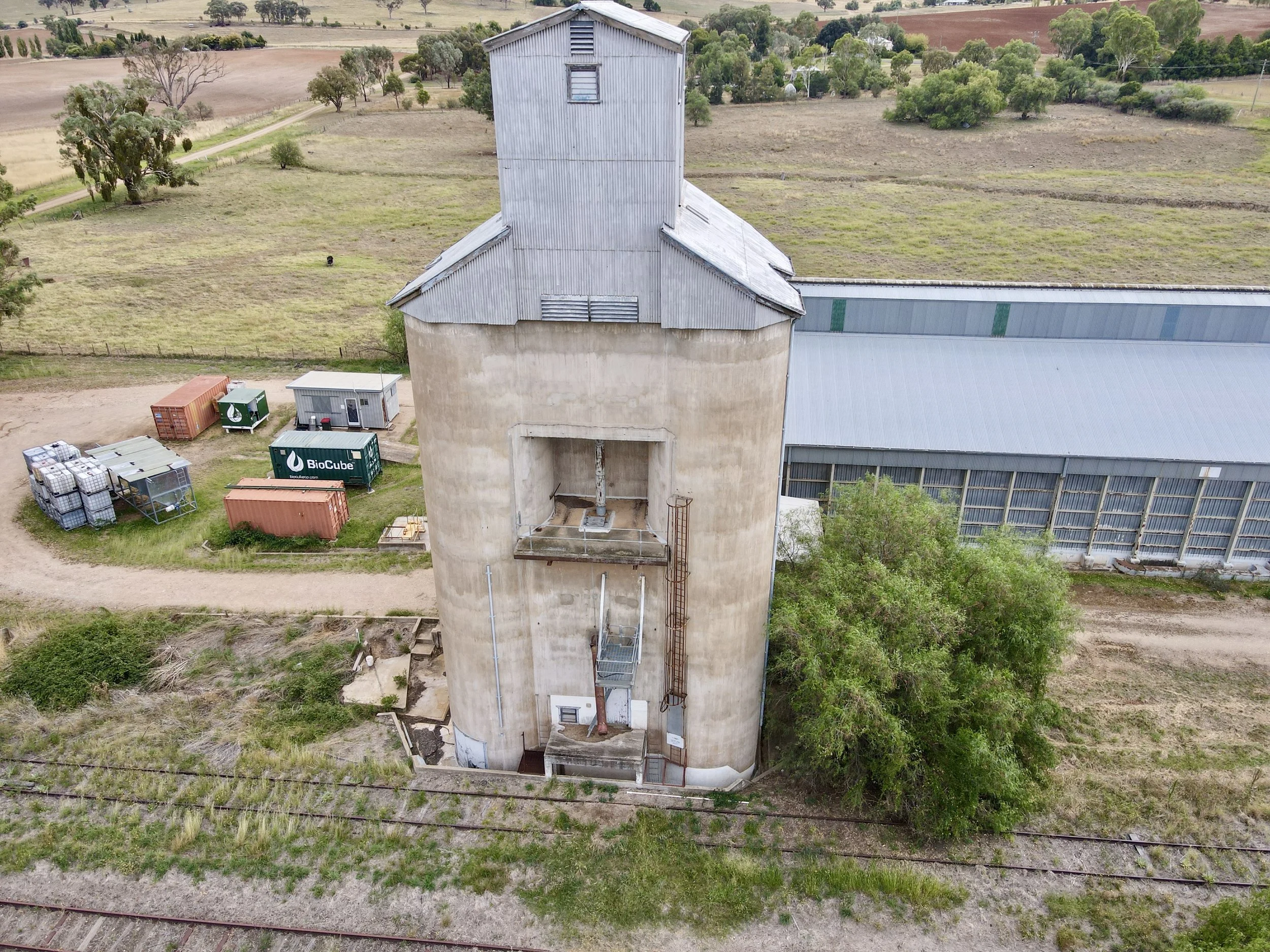 Aerial view of a cylindrical grain silo with a metal roof on a rural farm, surrounded by fields, trees, and farm structures.