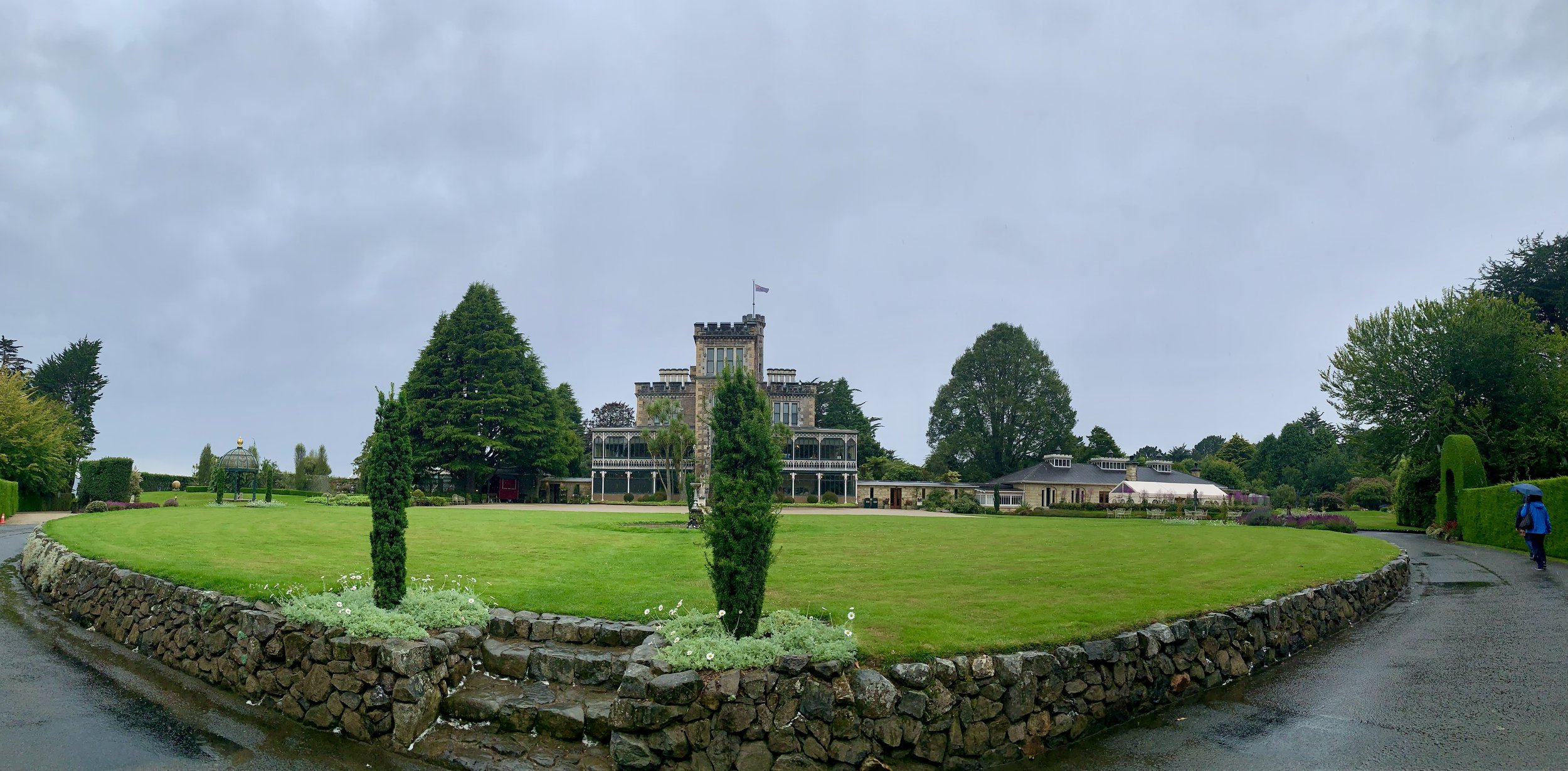 A large, stone castle-like mansion with multiple levels and a flag on top, surrounded by lush, green trees and manicured lawns on a cloudy day.