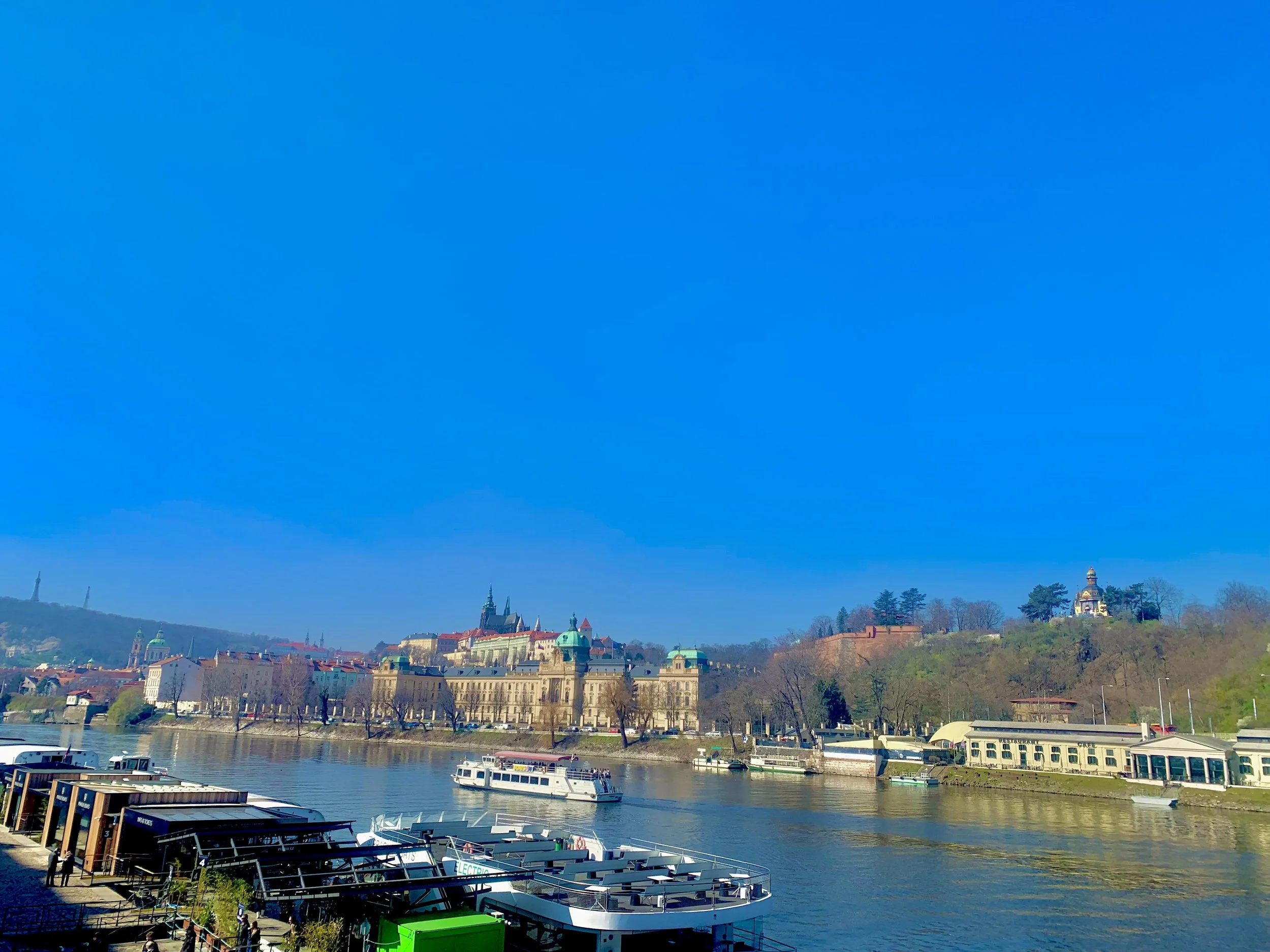 View of a river with boats docked at a pier in the foreground, historic buildings and a castle on a hill in the background, and a clear blue sky.