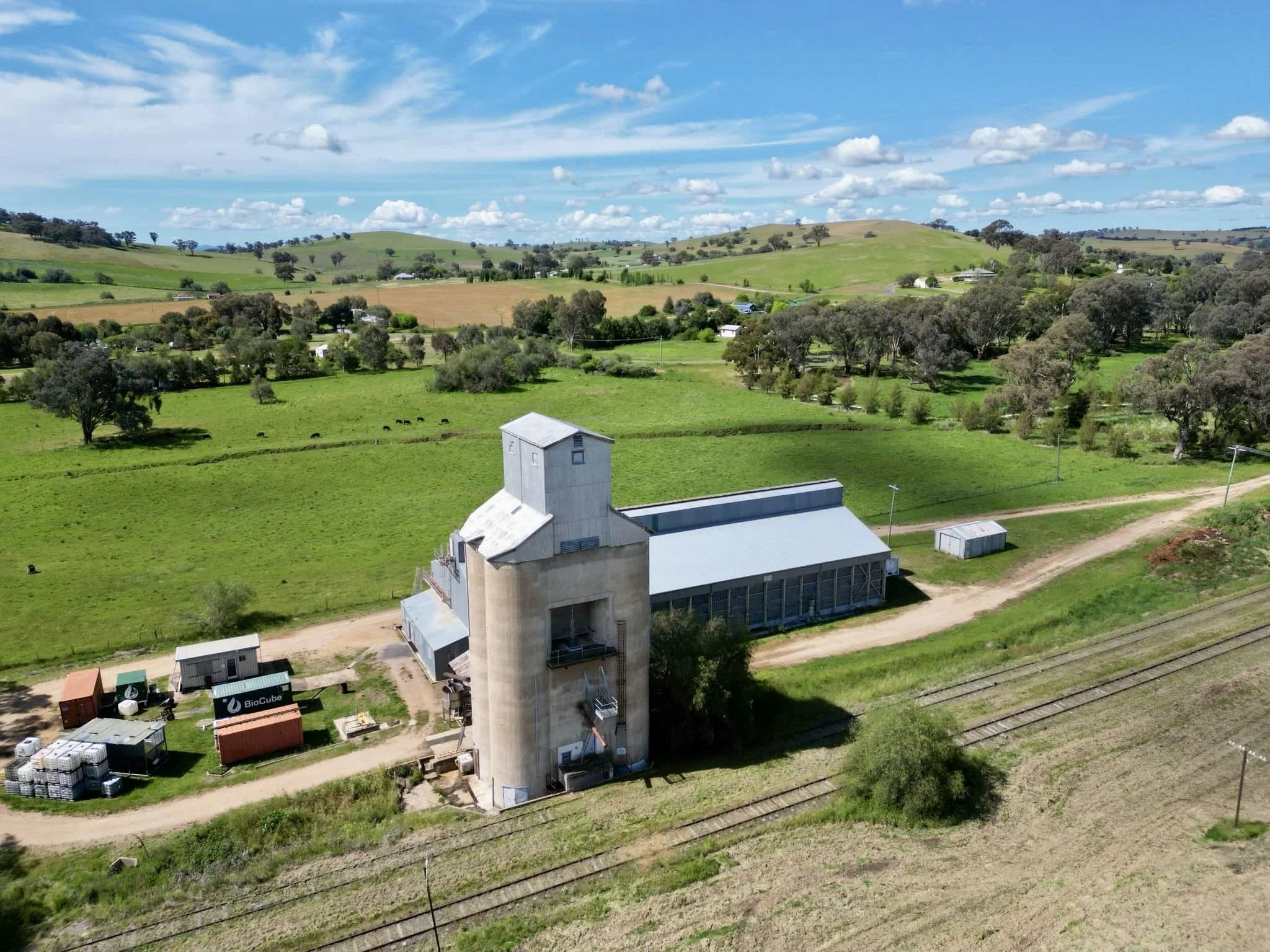 A rural landscape with a large silo and farm buildings surrounded by green fields, trees, and rolling hills under a blue sky with scattered clouds.