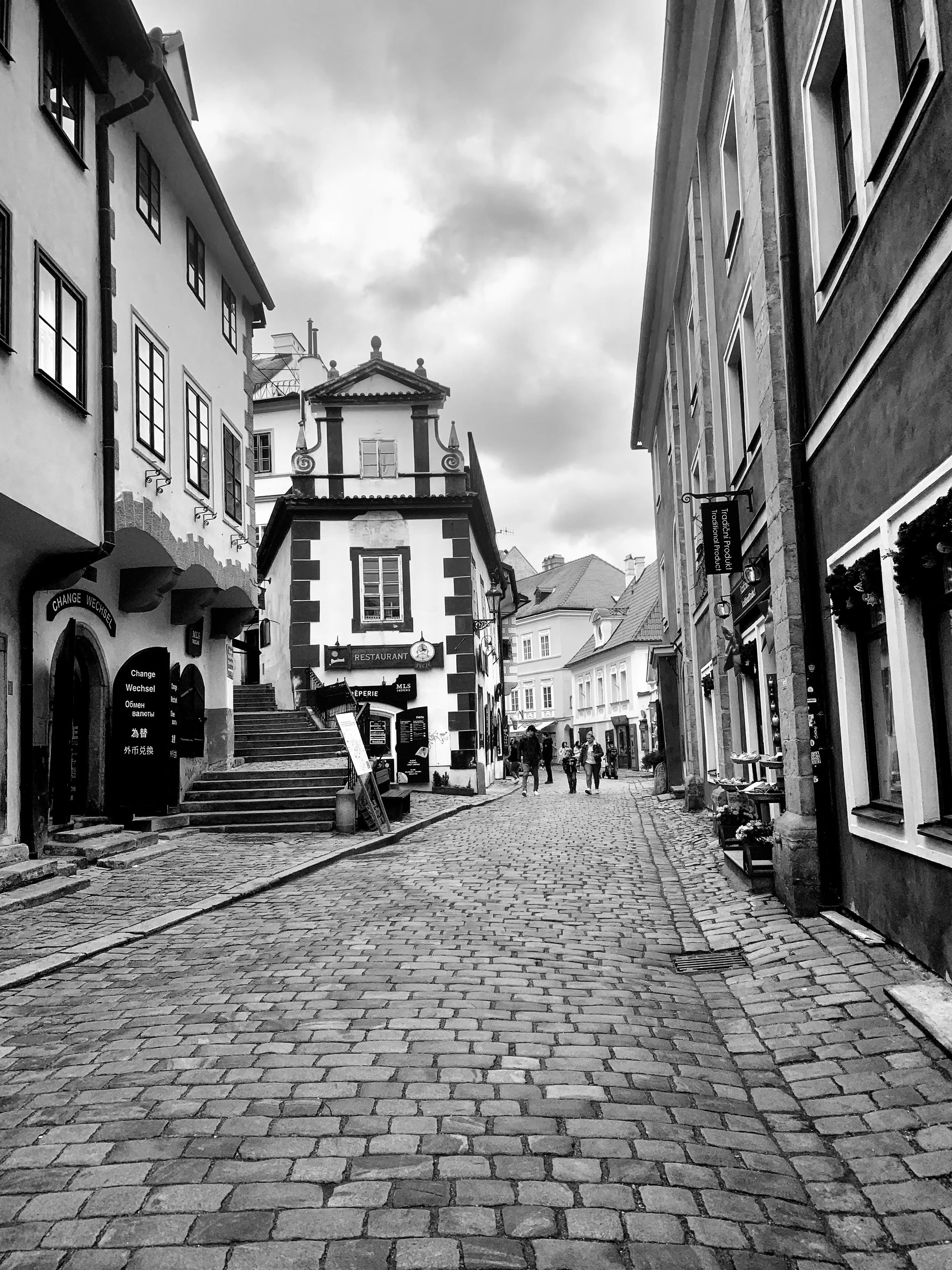 A cobblestone street in a European town with old buildings, some signs for restaurants, and a few people walking under a cloudy sky.