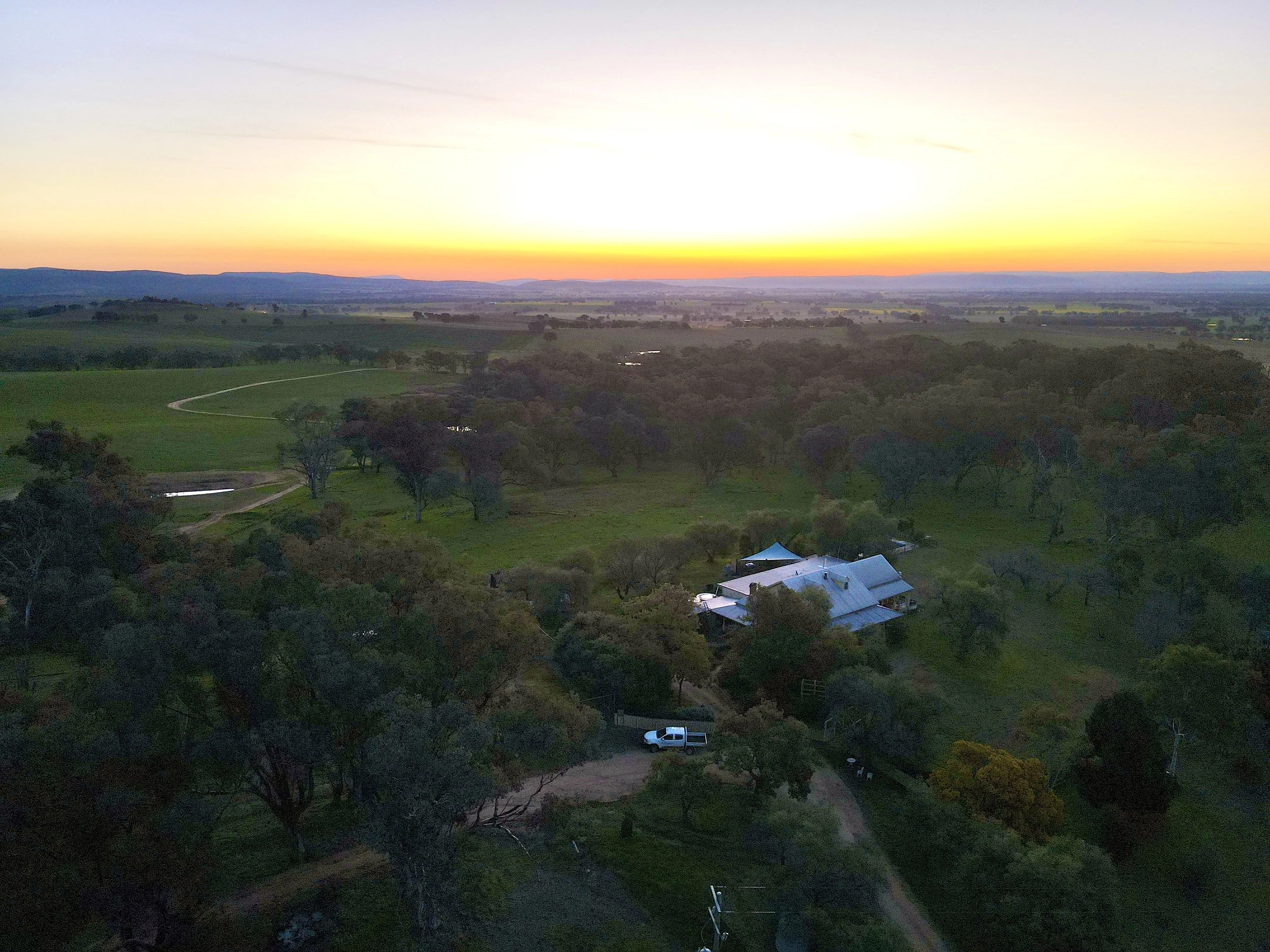Aerial view of a rural landscape at sunset with a farmhouse, trees, dirt road, and green fields.