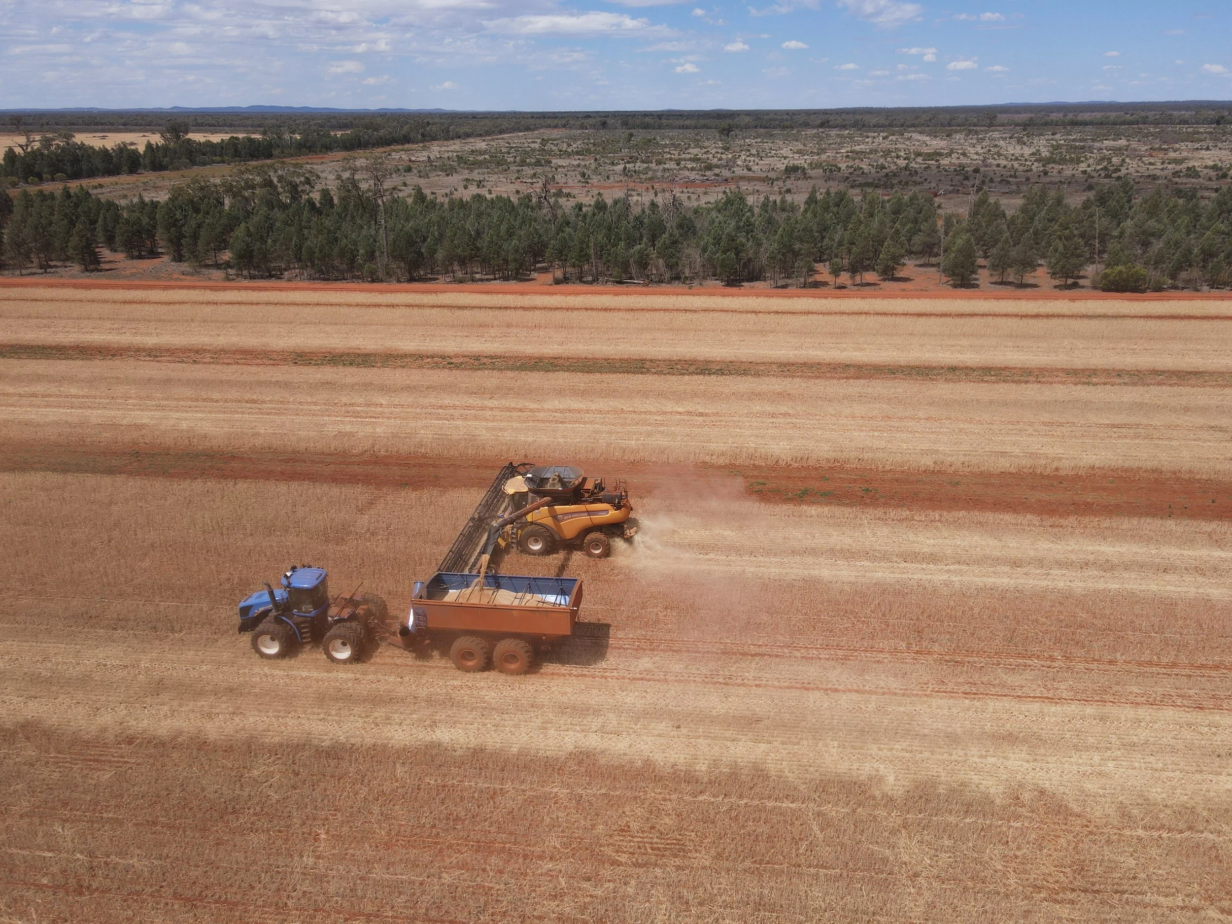 Two tractors and a harvester working on a large, open field of farmland, with a forested area in the background under a partly cloudy sky.