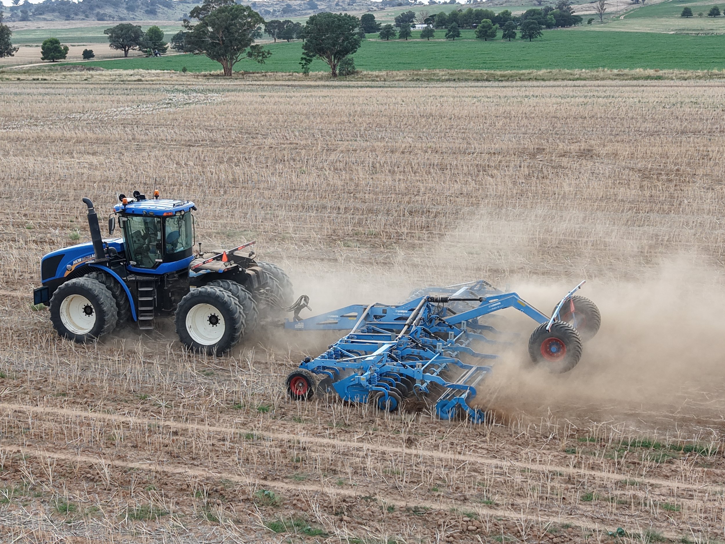 A blue tractor tilling a dry, harvested field with dusty soil in a rural landscape with trees and green fields in the background.