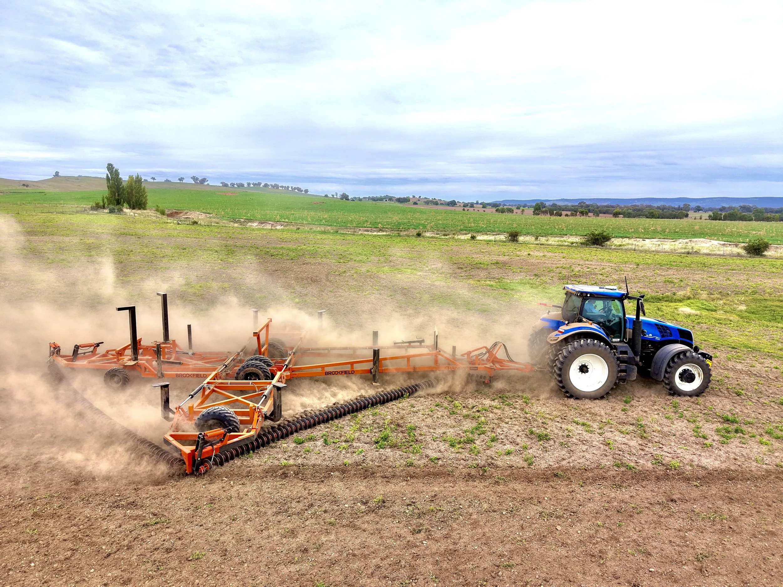 A blue tractor pulling an orange seed planter across a farm field, kicking up dust.
