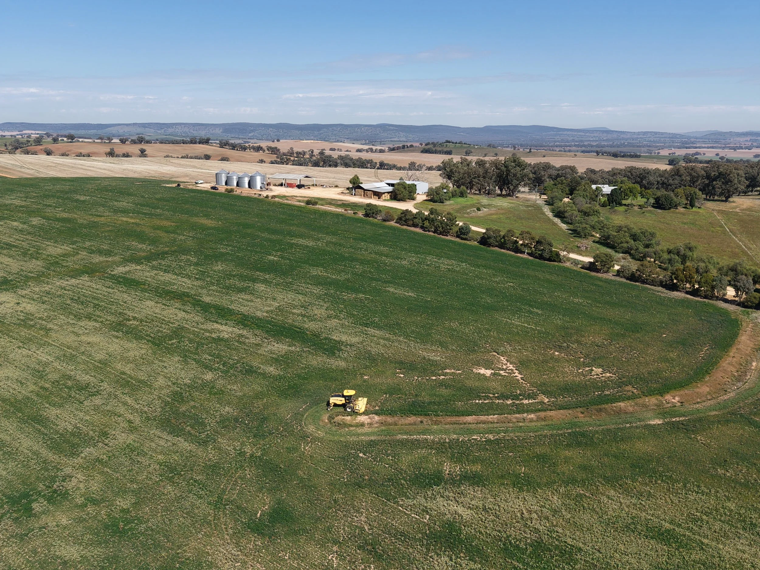 A yellow tractor working on a vast green farm field with a dirt path, farmland, silos, farm buildings, trees, and rolling hills in the distance.