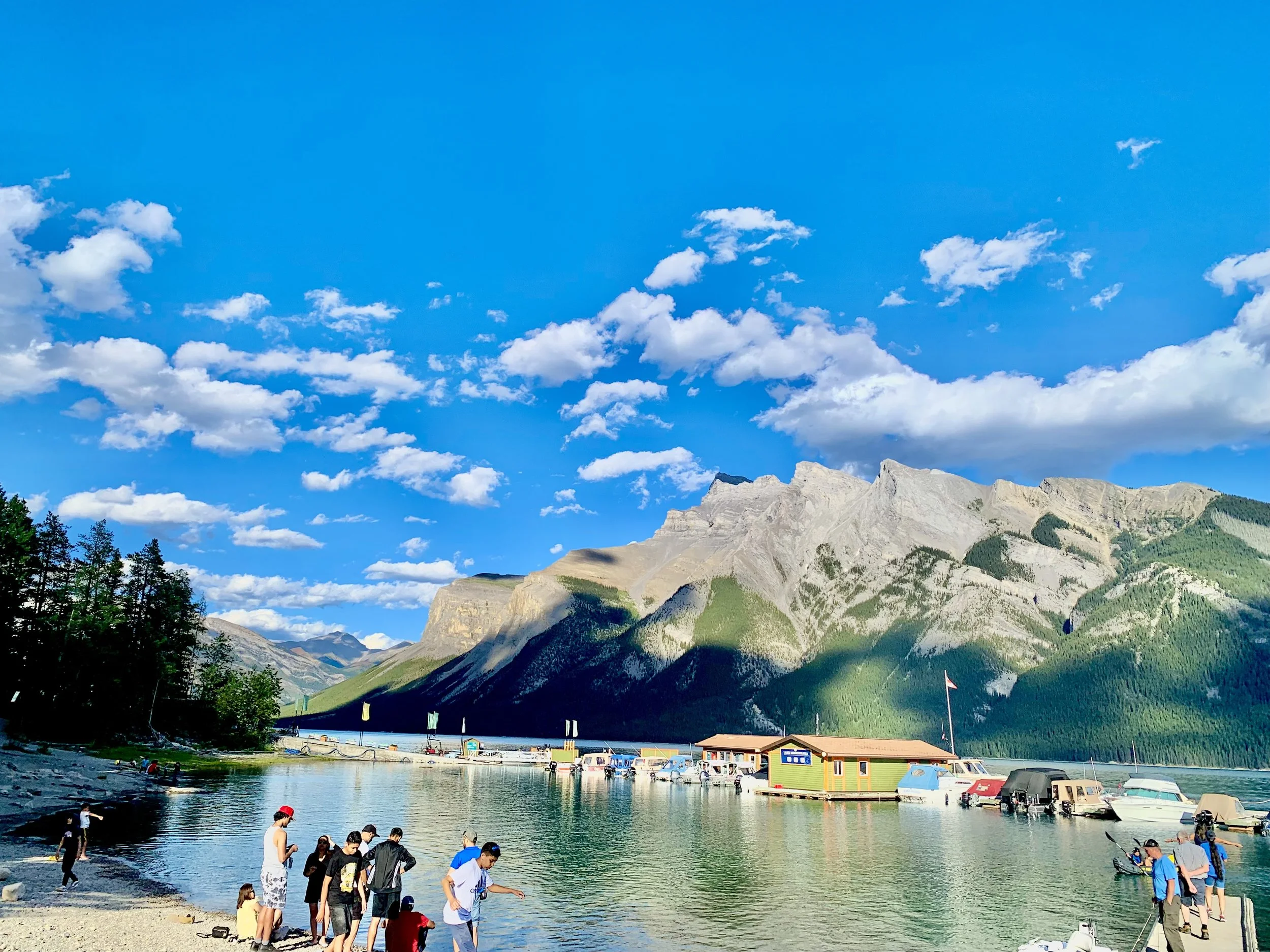 People gathered by a lake surrounded by mountains and trees, with boats docked at a floating dock, under a partly cloudy blue sky.