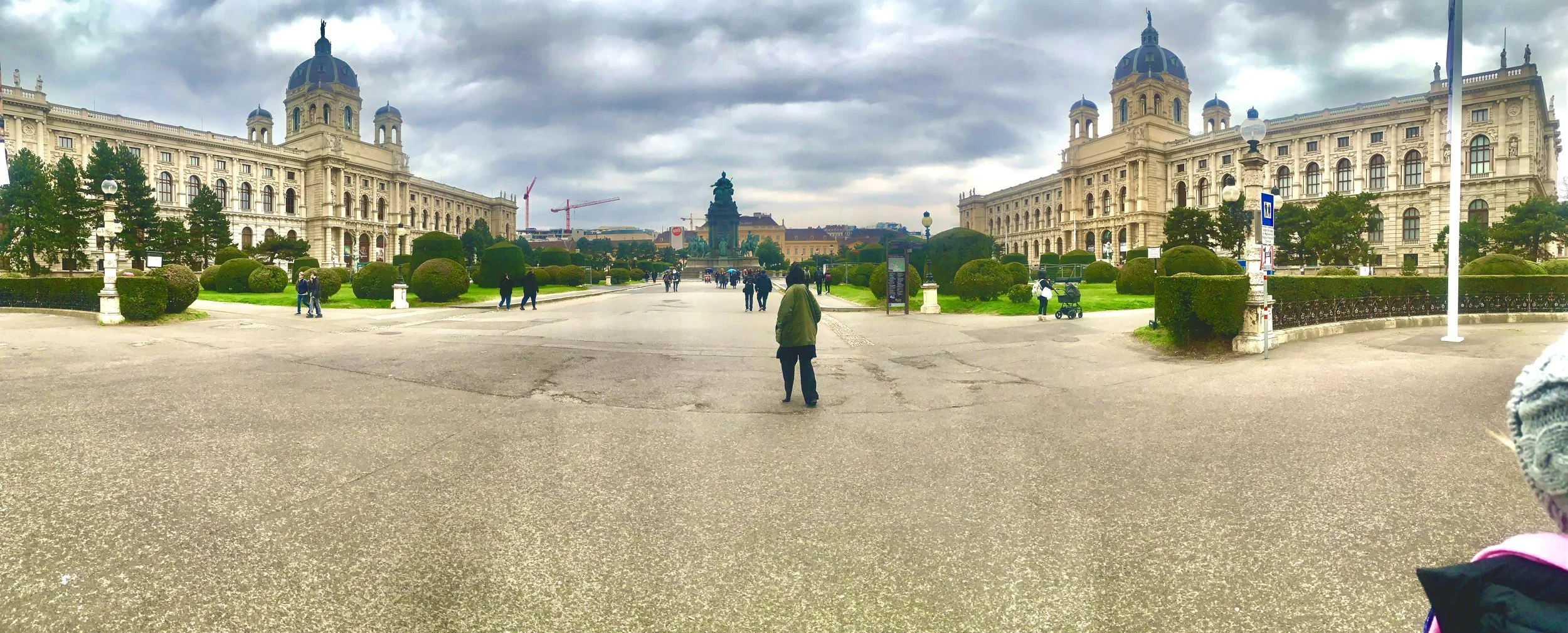 A wide view of a historic building with domed towers, a central square with people walking, green shrubs, and cloudy sky.