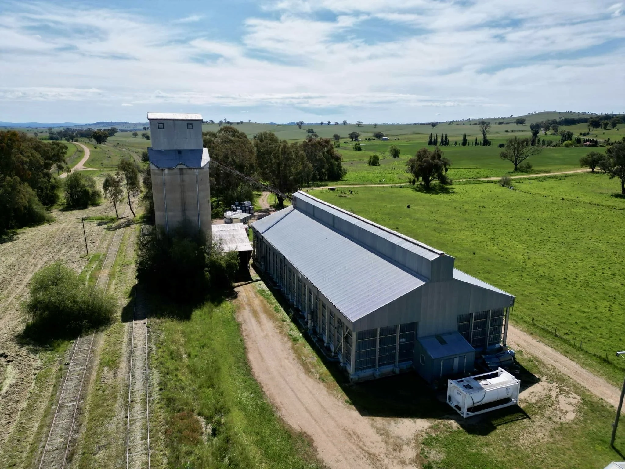 Aerial view of a rural farm with a large metal barn, a tall grain silo, train tracks, green fields, and a dirt road with scattered trees extending into the distance under a partly cloudy sky.