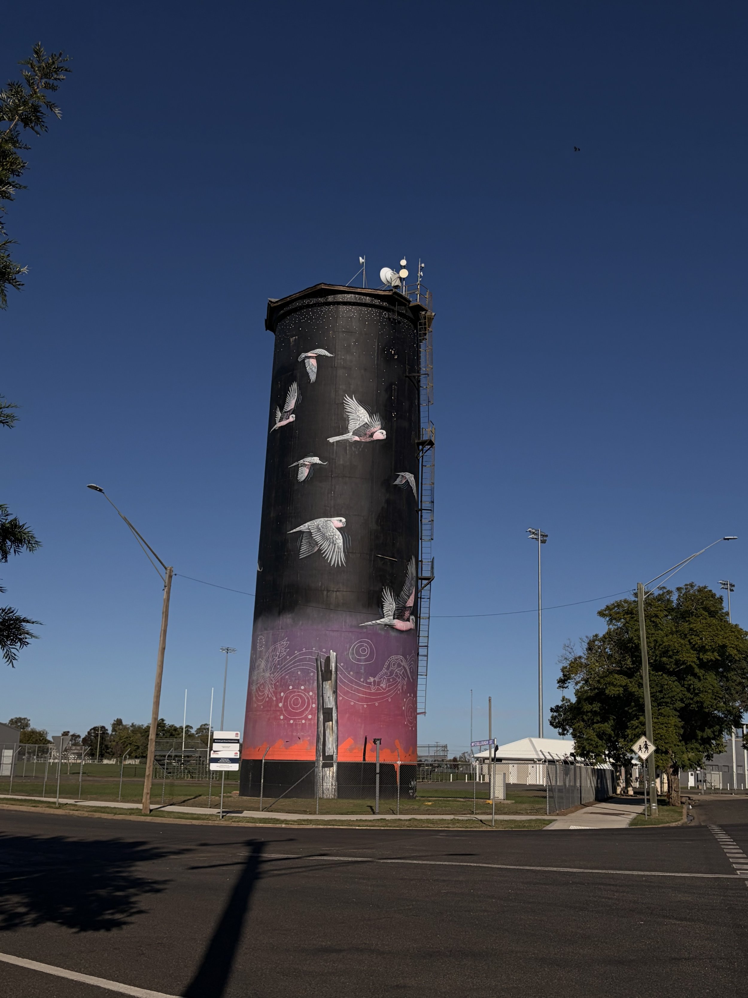 A tall black tower with painted birds, including pink and gray tones, set against a clear blue sky with some trees and a fence in the foreground.