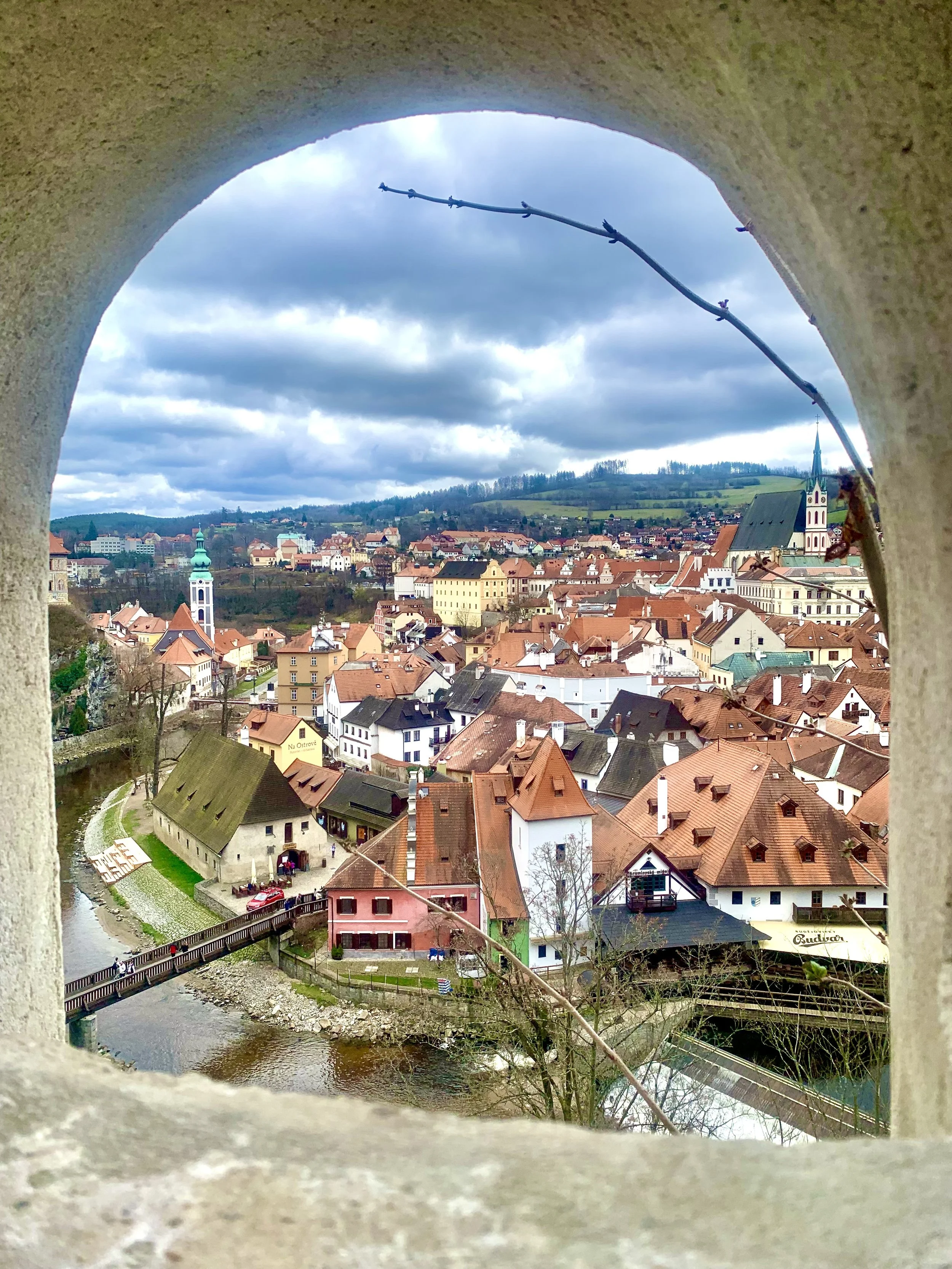 View of a European town with colorful buildings, church steeples, a river, and rolling hills, seen through a circular opening in a stone wall.