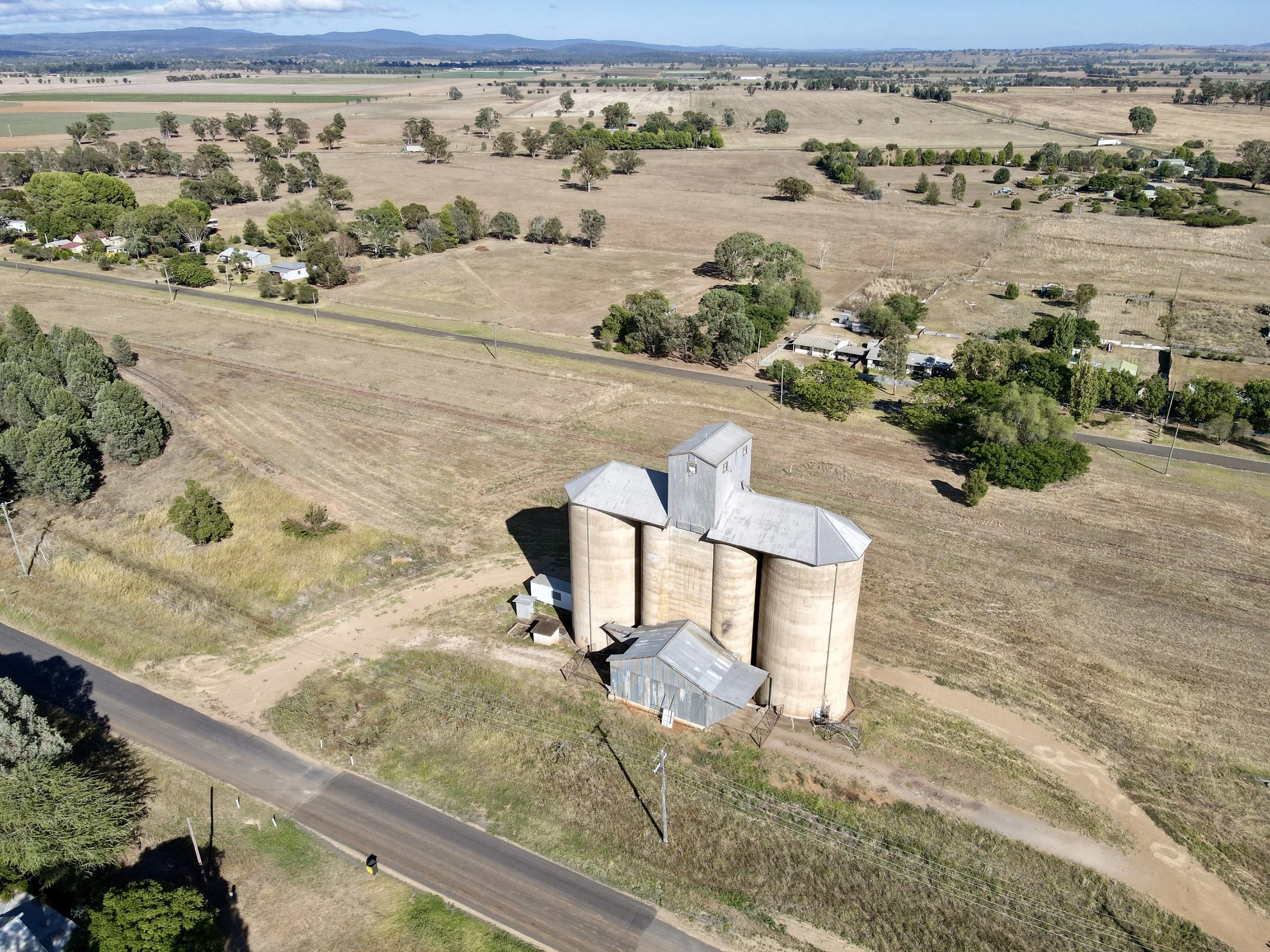An aerial view of a rural landscape with a small cluster of farm buildings and silos in the foreground, surrounded by fields, trees, and a few scattered houses.