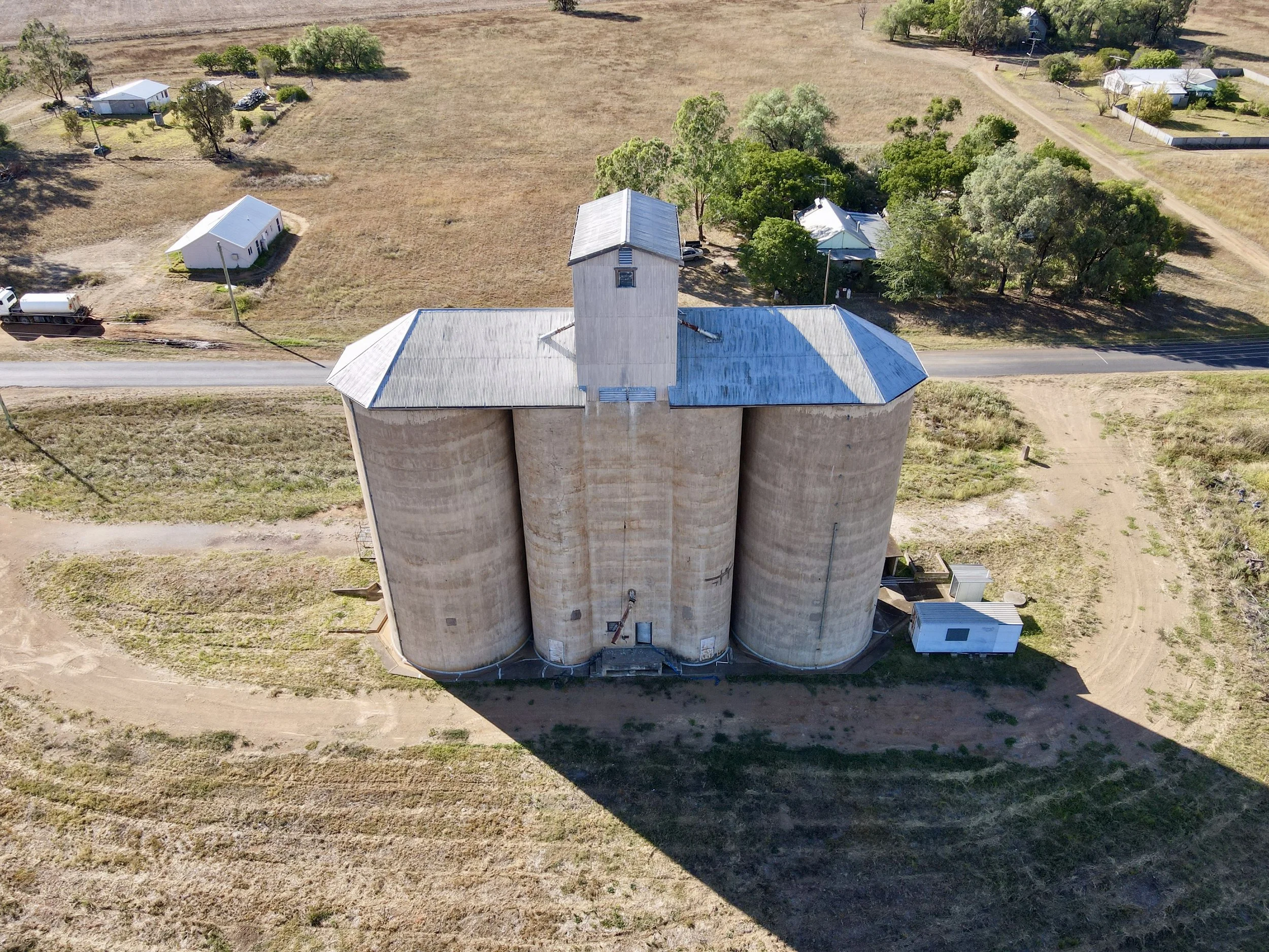 Aerial view of a large beige grain silo with a metallic roof, casting a long shadow on the ground, situated near a rural road with a few small buildings and trees around.
