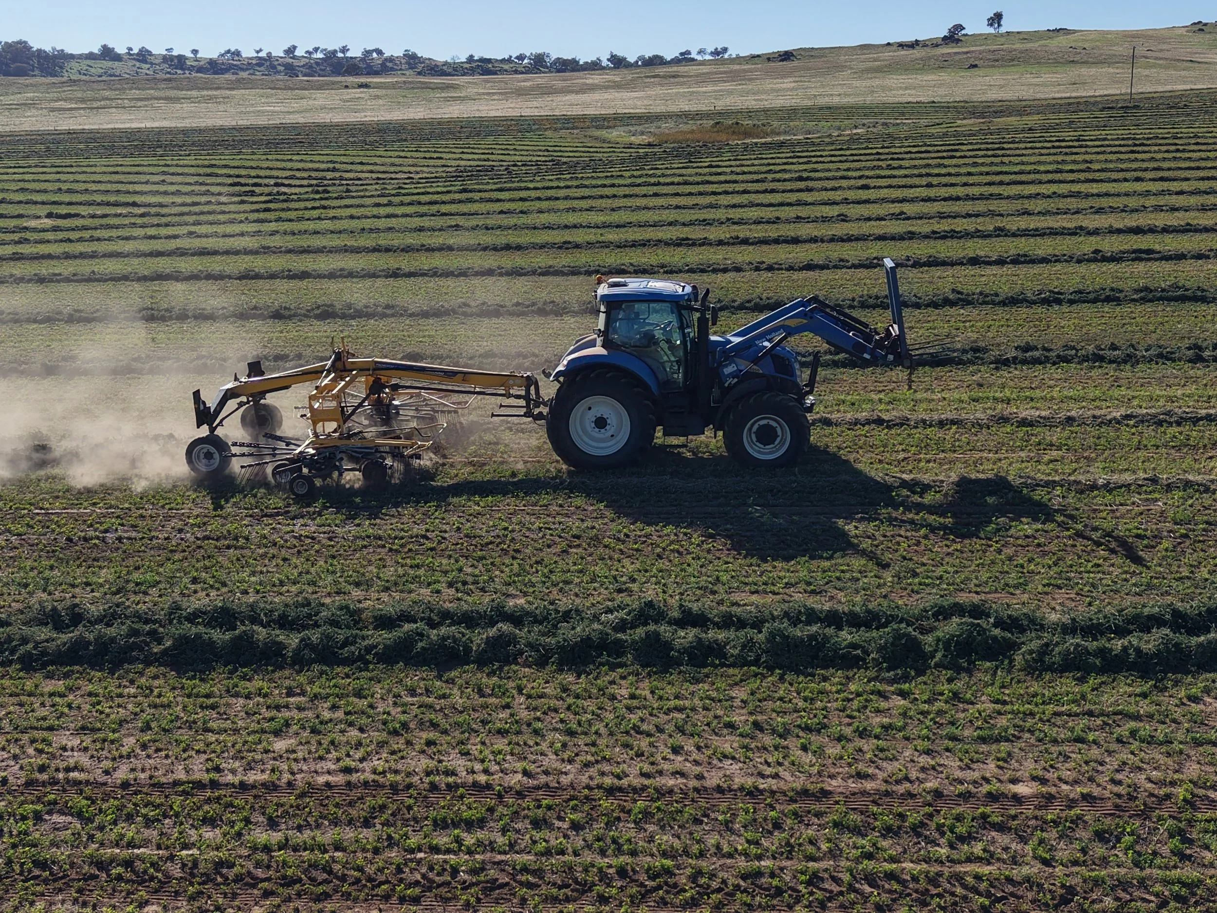 A tractor is plowing a field, kicking up dust as it moves across the farmland with multiple rows of crops, hills, and trees in the background.