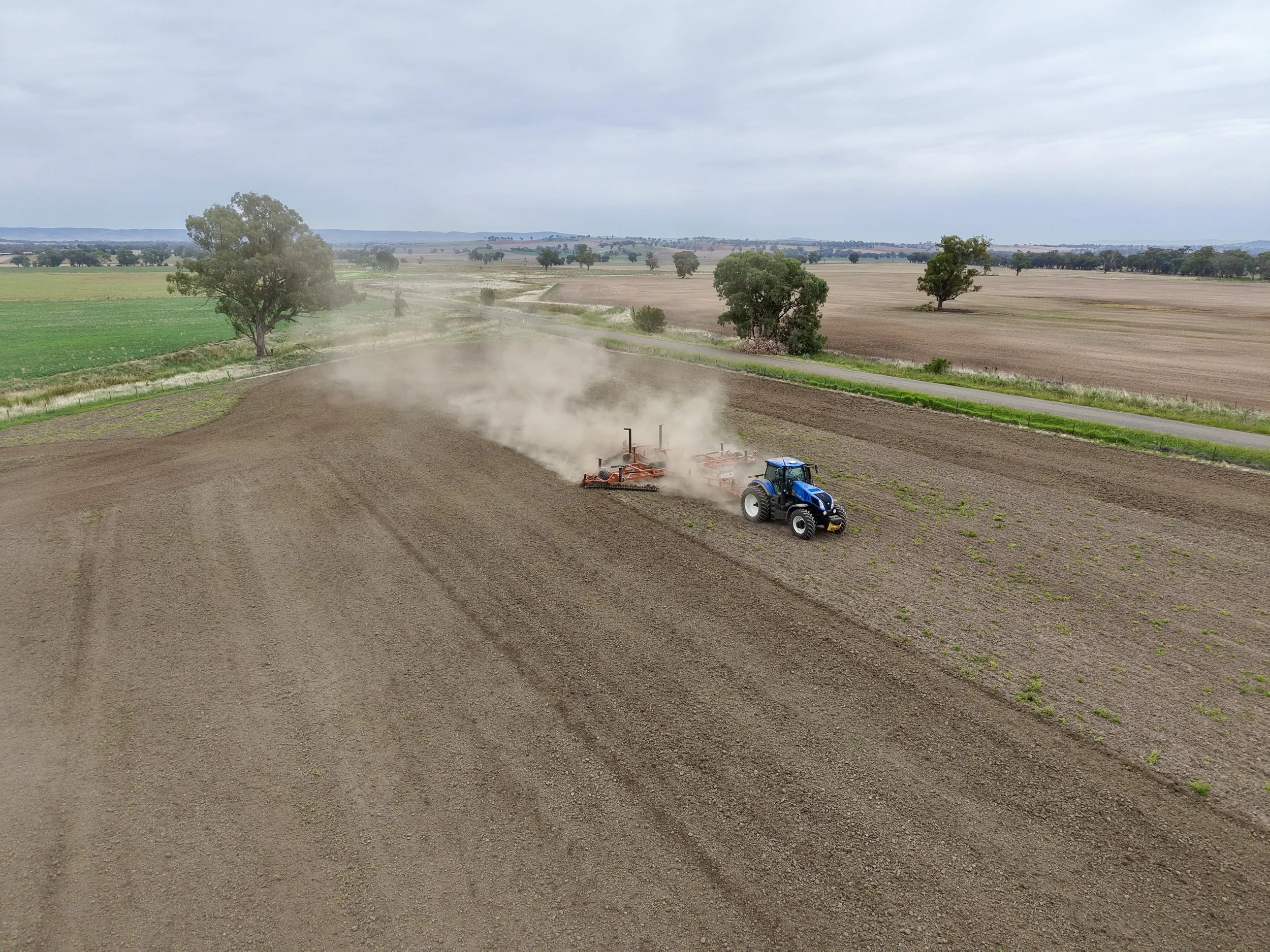 A blue tractor pulling farming equipment in a large, open field, with dust rising from the soil as it works the land, under a cloudy sky.