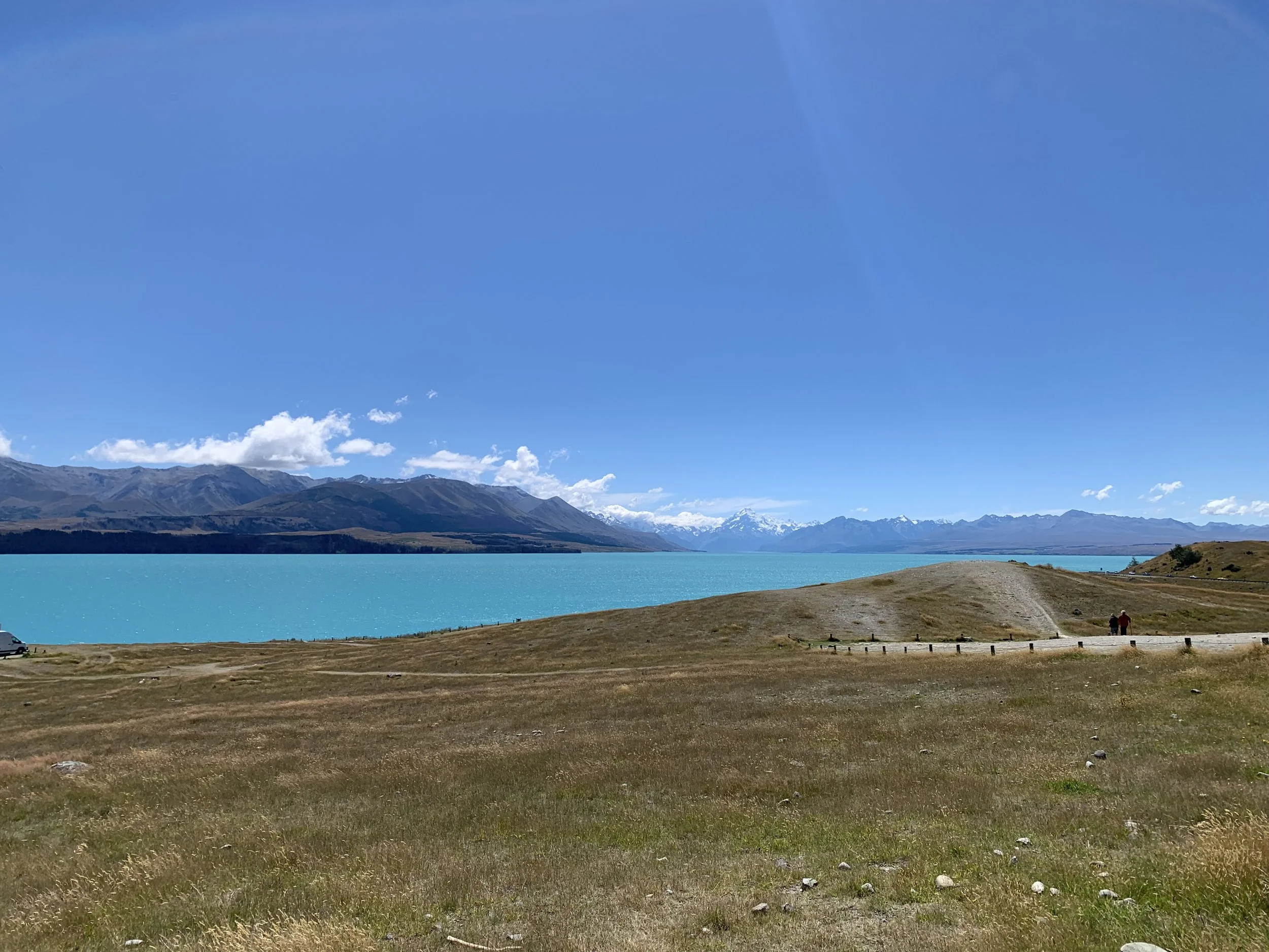 A landscape with a bright blue lake, distant snowy mountains, a grassy foreground, and a clear blue sky.