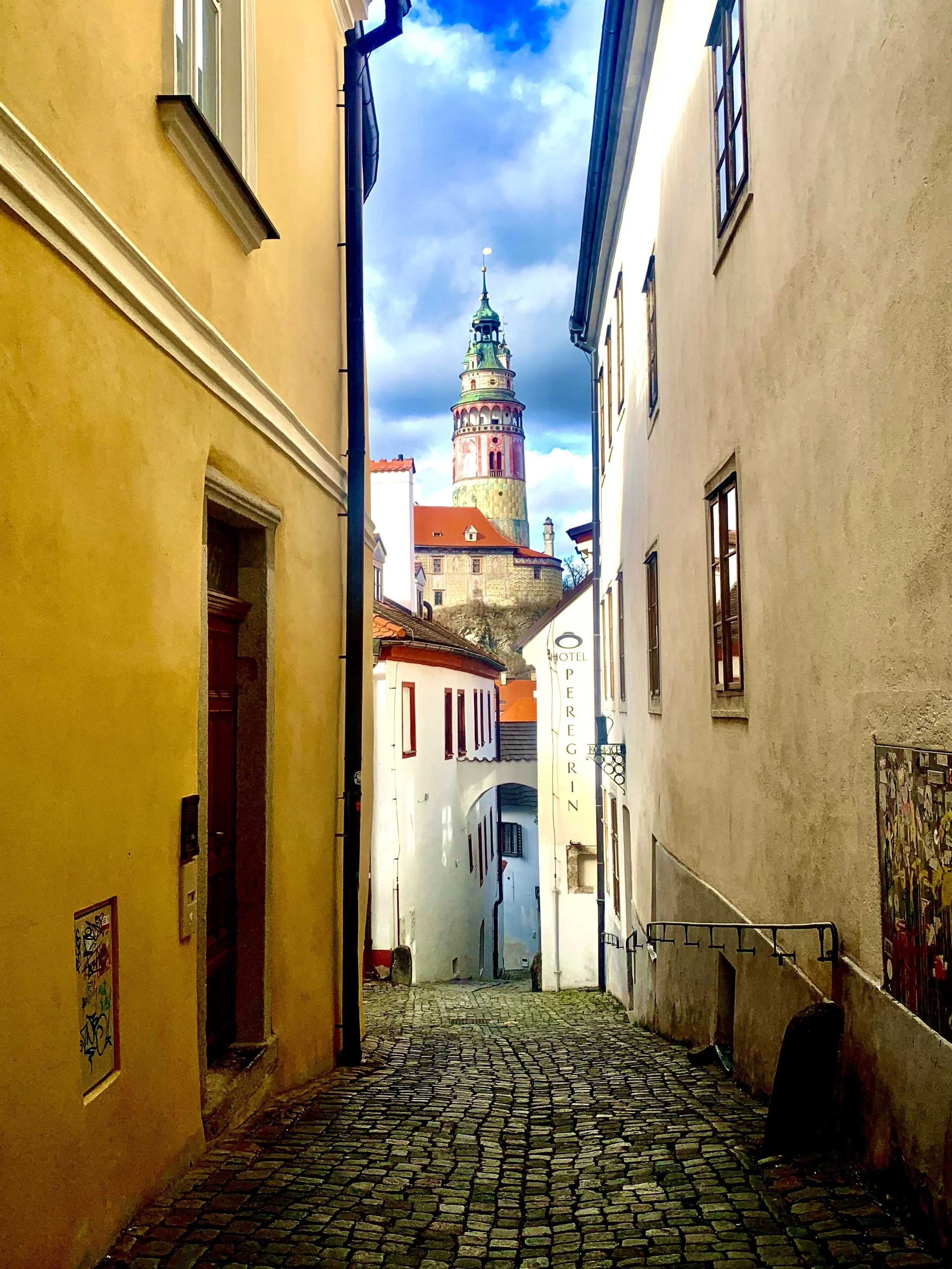 A narrow cobblestone street between yellow and beige buildings, leading to a view of a colorful castle tower against a partly cloudy sky.