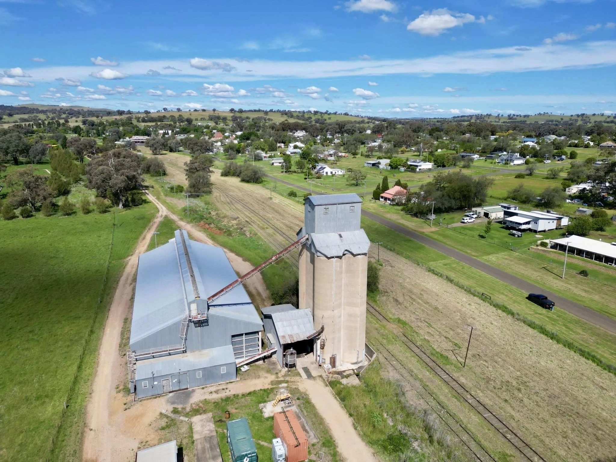 An aerial view of a rural farm with a grain silo and storage buildings, fields, and nearby residential houses under a partly cloudy sky.
