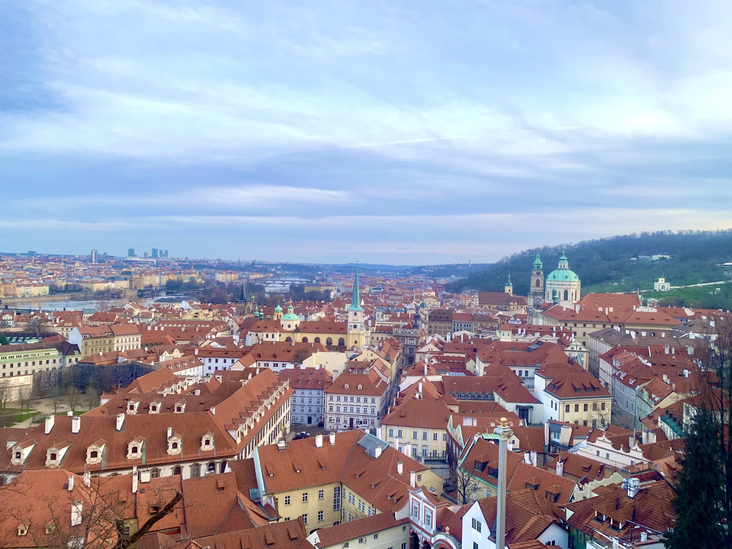 A scenic view of a European city with red-tiled rooftops, church steeples, and hills in the background under a partly cloudy sky.