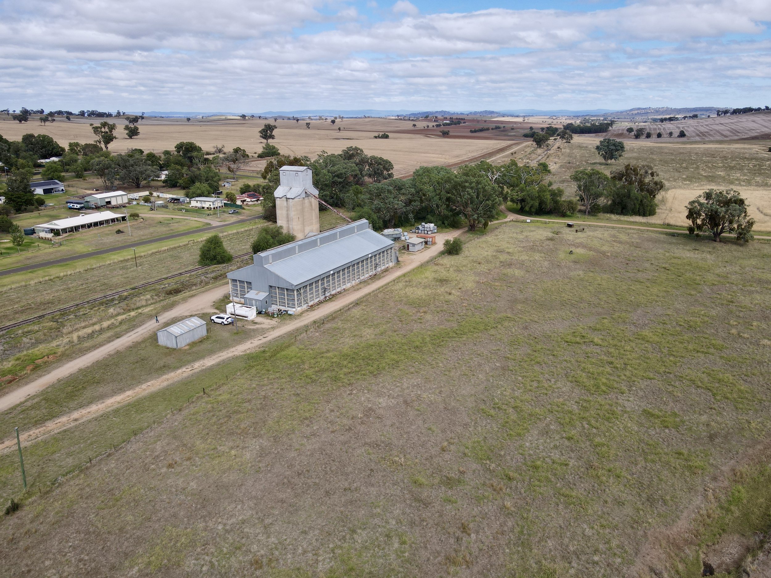 An aerial view of a rural farm with a large barn, silo, and surrounding fields, with empty land and scattered trees in the background under a partly cloudy sky.
