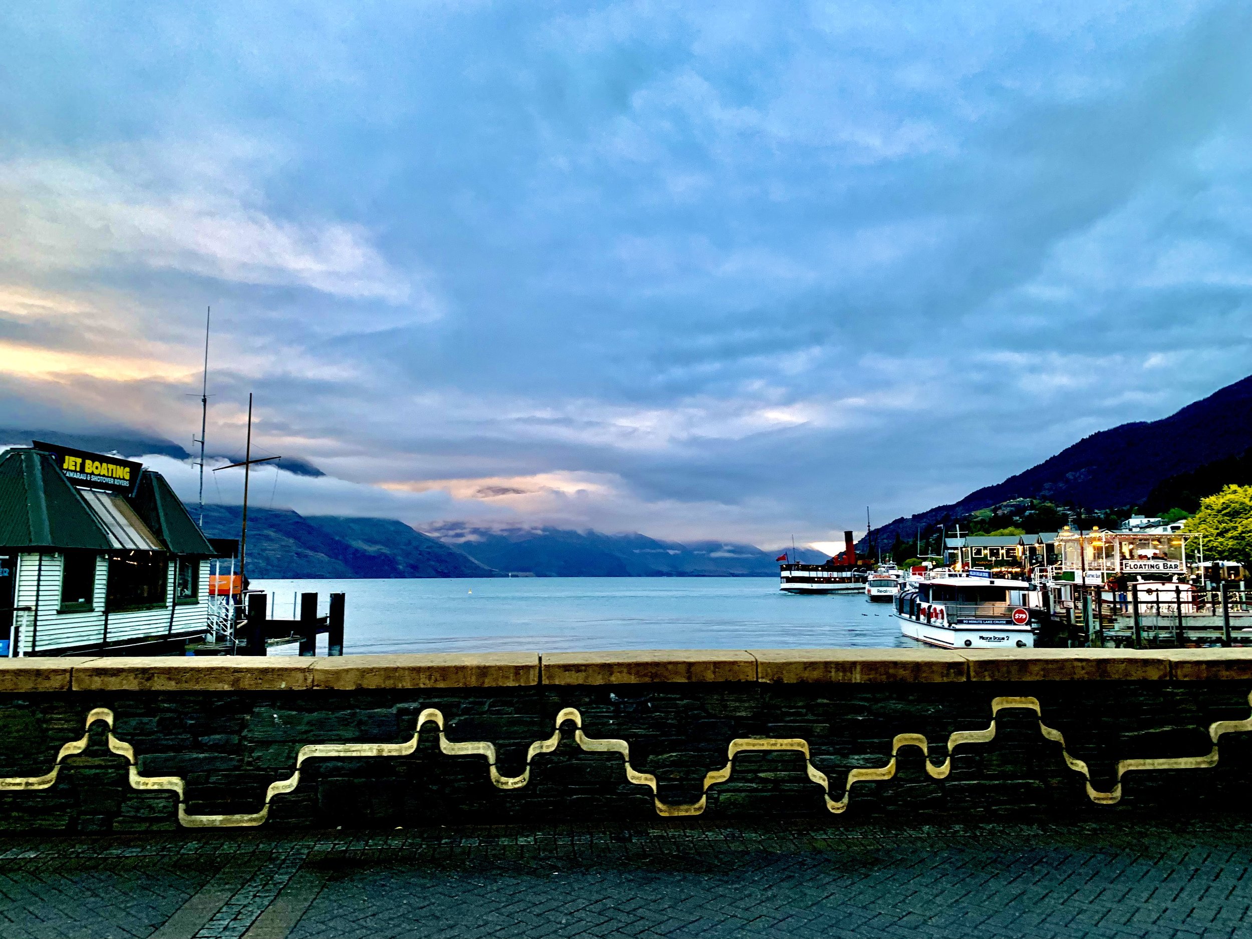 Marina with docked boats, mountain backdrop, cloudy sky, and a small building with a sign that says 'Jet Boating'.